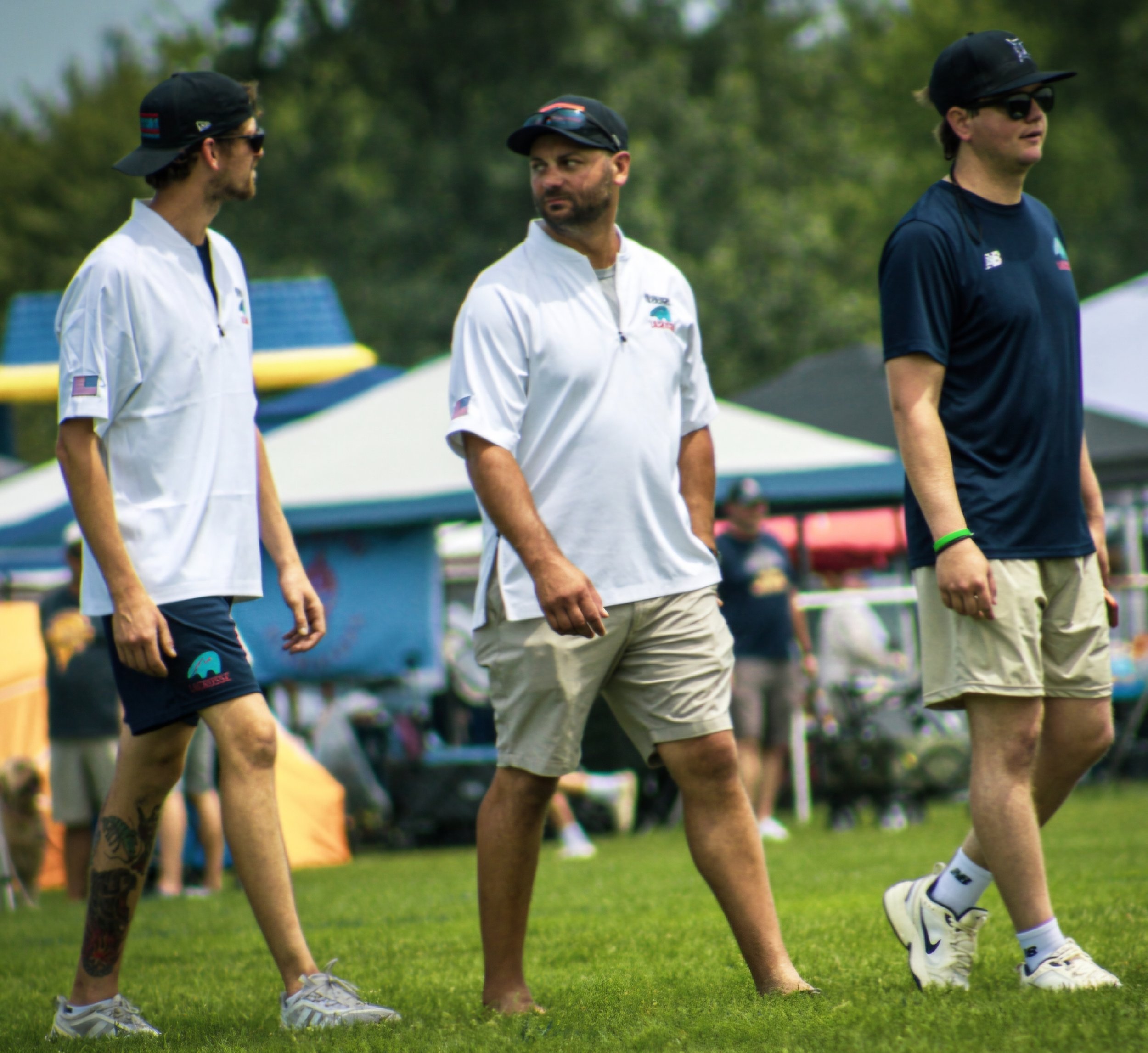 Three men walking in a line outdoors on grass, wearing casual sports clothing including shorts, t-shirts, and caps, with trees and a tent or canopy in the background.