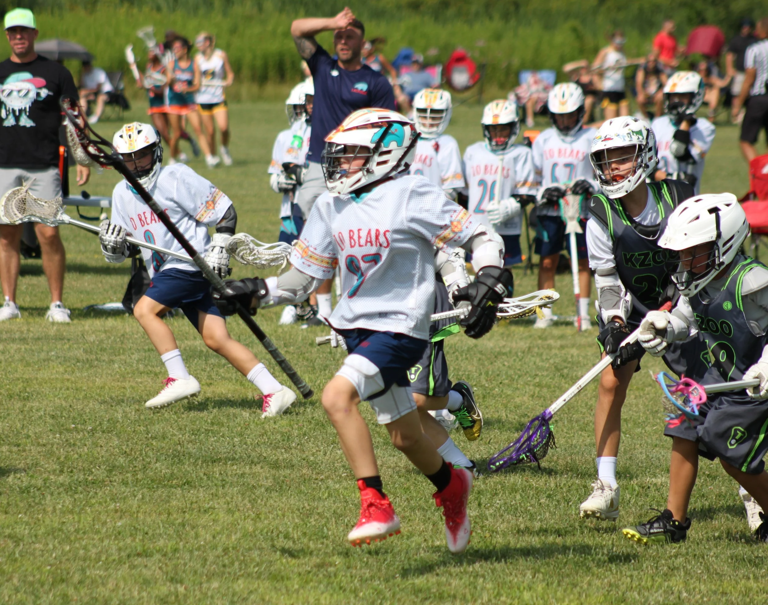Kids playing lacrosse on a grassy field with spectators and coaches in the background during a sunny day.