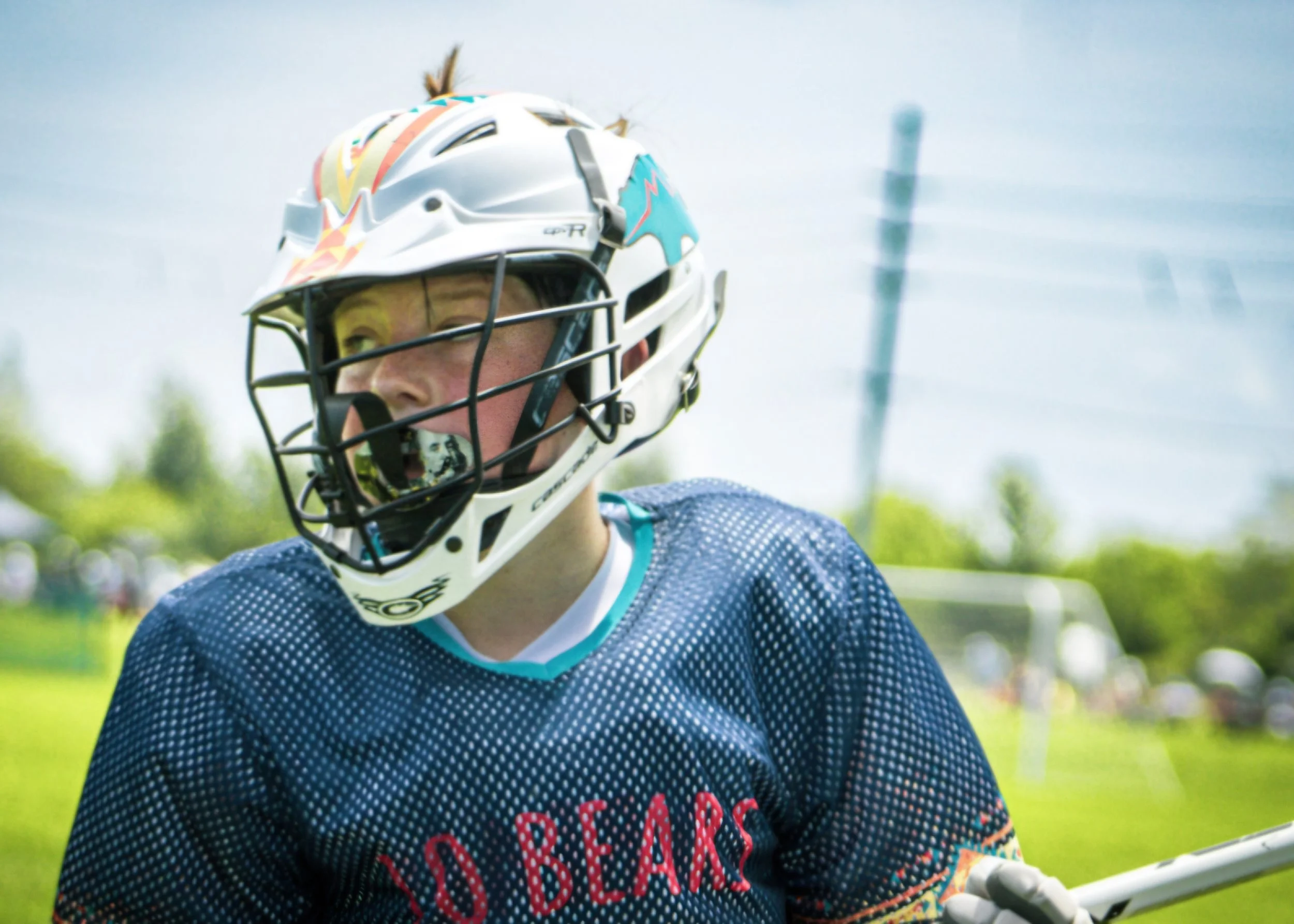 Close-up of a young athlete wearing a football helmet with a faceguard, and a blue jersey with red and white lettering, outdoors on a sunny day.