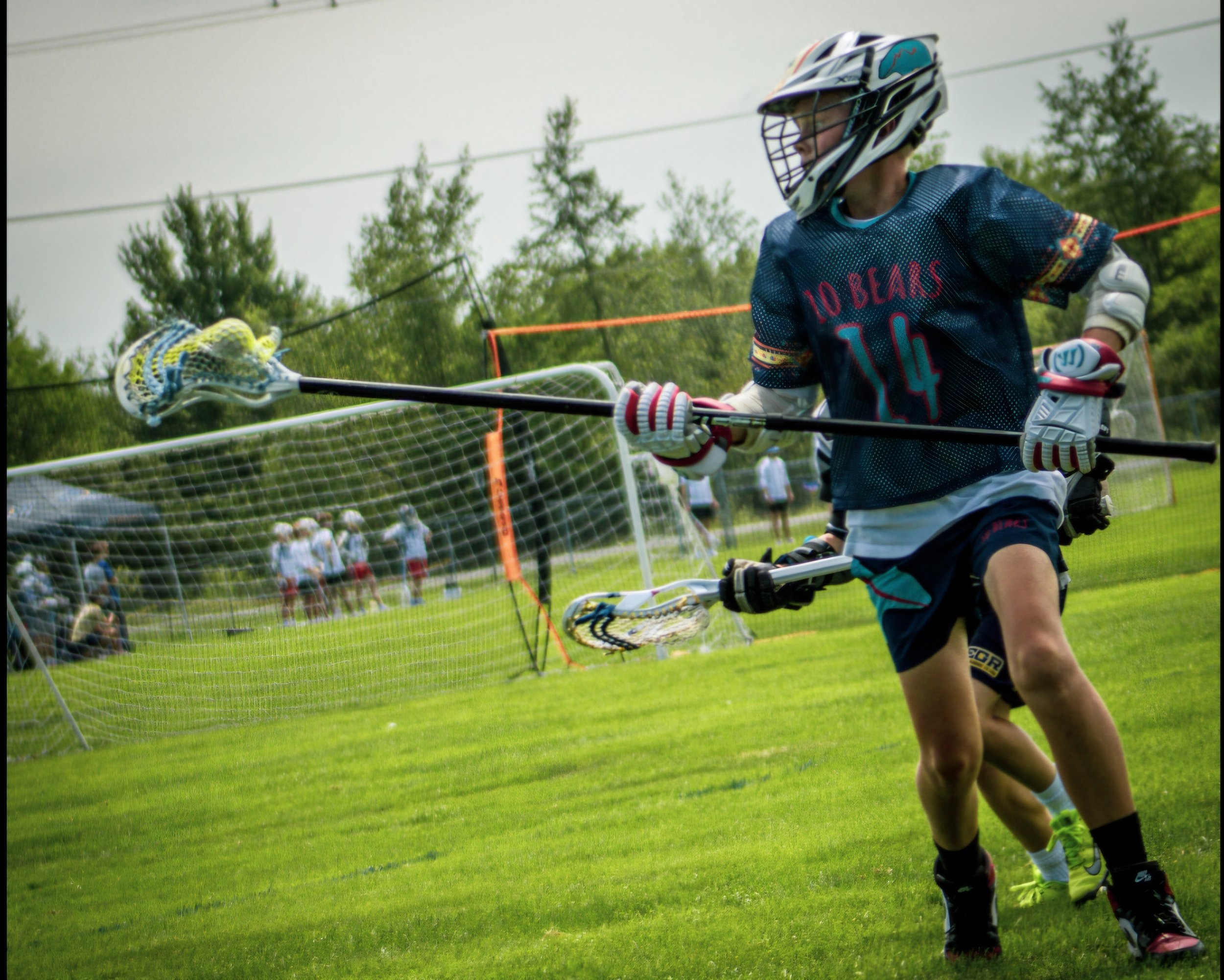 Lacrosse player wearing blue jersey with red lettering, helmet with face guard, and gloves holding a lacrosse stick in action on the field during a game, with a net and spectators in the background.