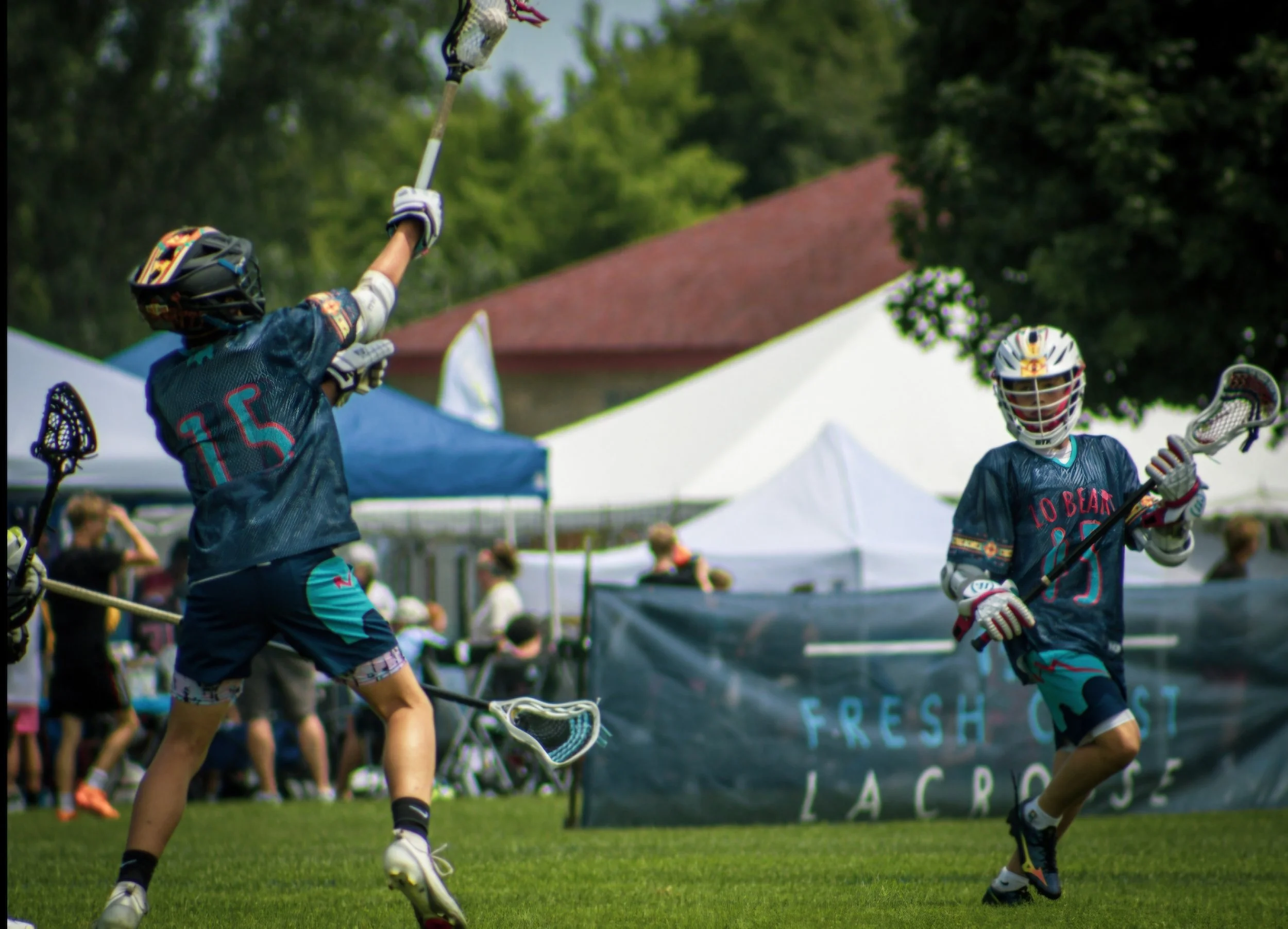 Two young male lacrosse players wearing helmets, gloves, and jerseys playing on a grassy field during a game. One player is reaching up with his lacrosse stick, and the other is holding his lacrosse stick while looking at the ball. Tents and spectato