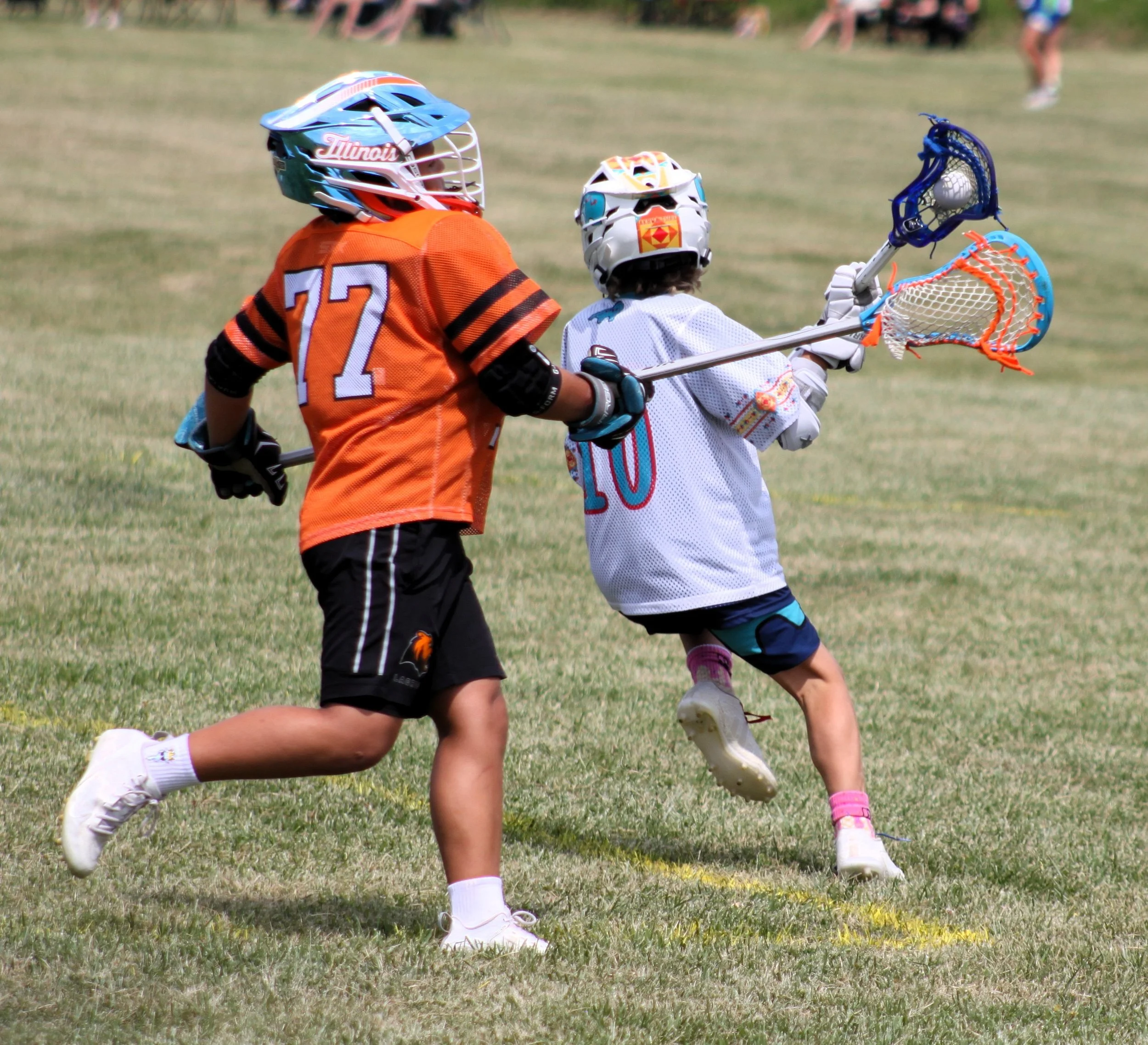 Two young kids playing lacrosse on a grassy field, wearing helmets and sports jerseys, with the back view visible and the field extending into the background.