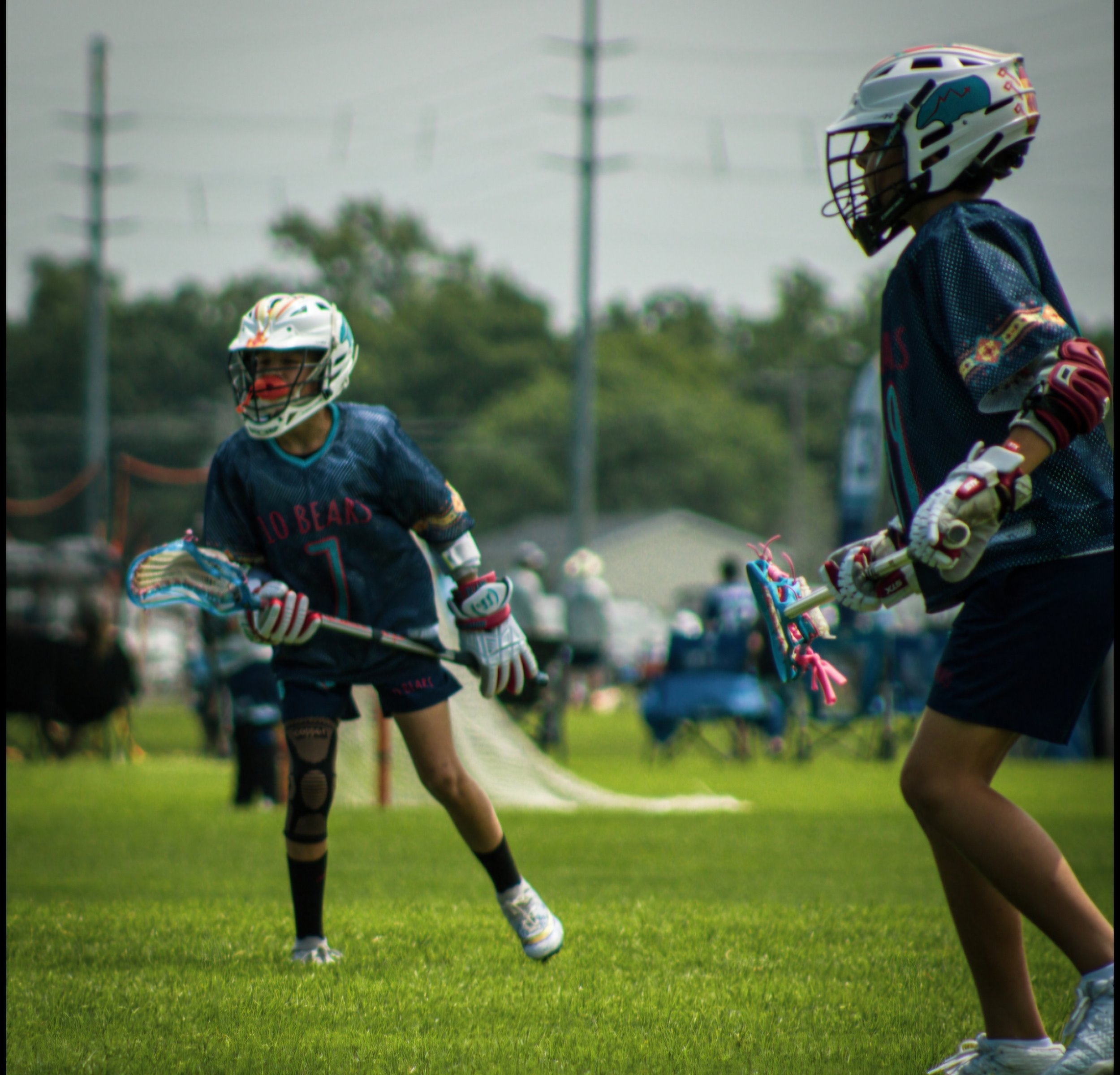 Two young lacrosse players in sports gear and helmets, actively engaged in a game on a grassy field.