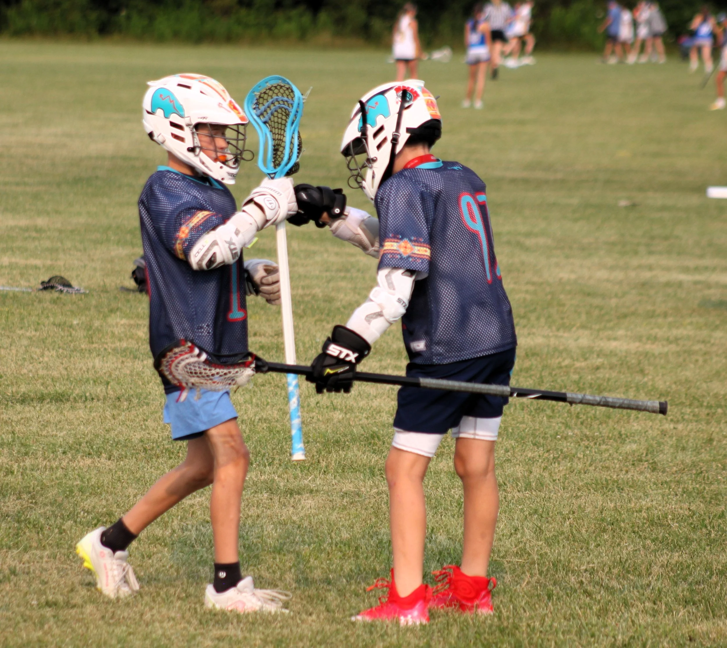 Two young boys wearing helmets and sports gear shaking hands on a grassy field during a youth lacrosse game, with a group of girls in the background.