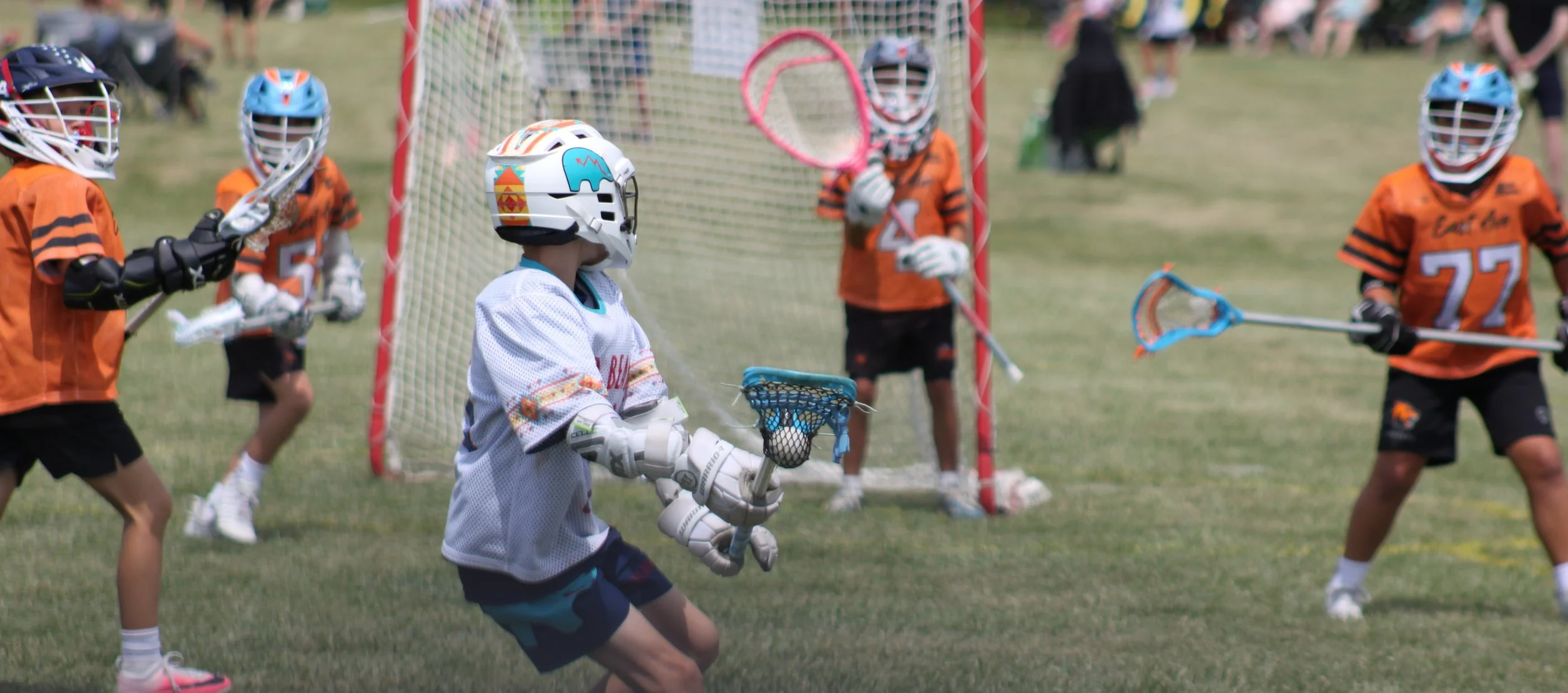 Children playing lacrosse on a grassy field, with one child in white gear and others in orange jerseys, some wearing helmets and holding lacrosse sticks, near a goal net.