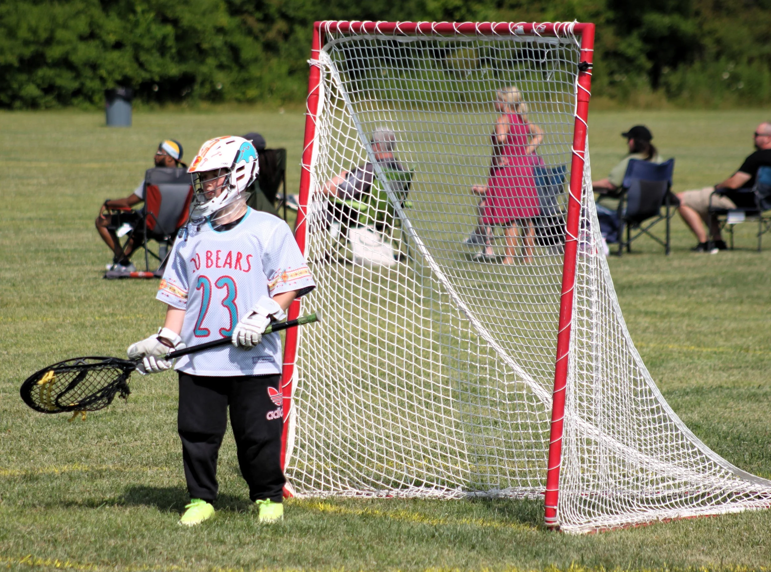 A young girl dressed in a hockey jersey, gloves, and helmet holding a hockey stick stands next to a soccer goal on a grassy field, with spectators sitting in chairs in the background.