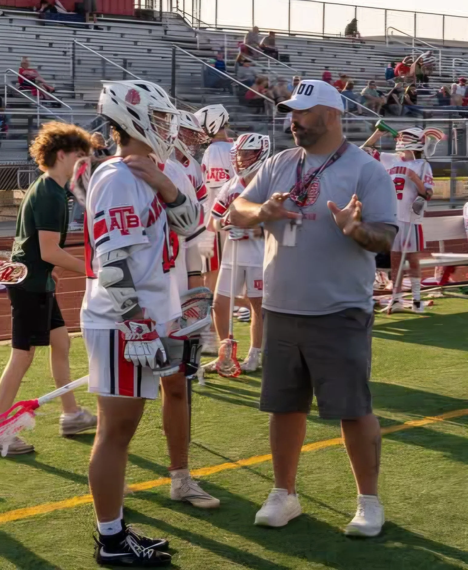 A group of lacrosse players and a coach standing on a football field, engaged in conversation during a game or practice, with players wearing white uniforms and helmets, and one person in casual clothing with a white cap.