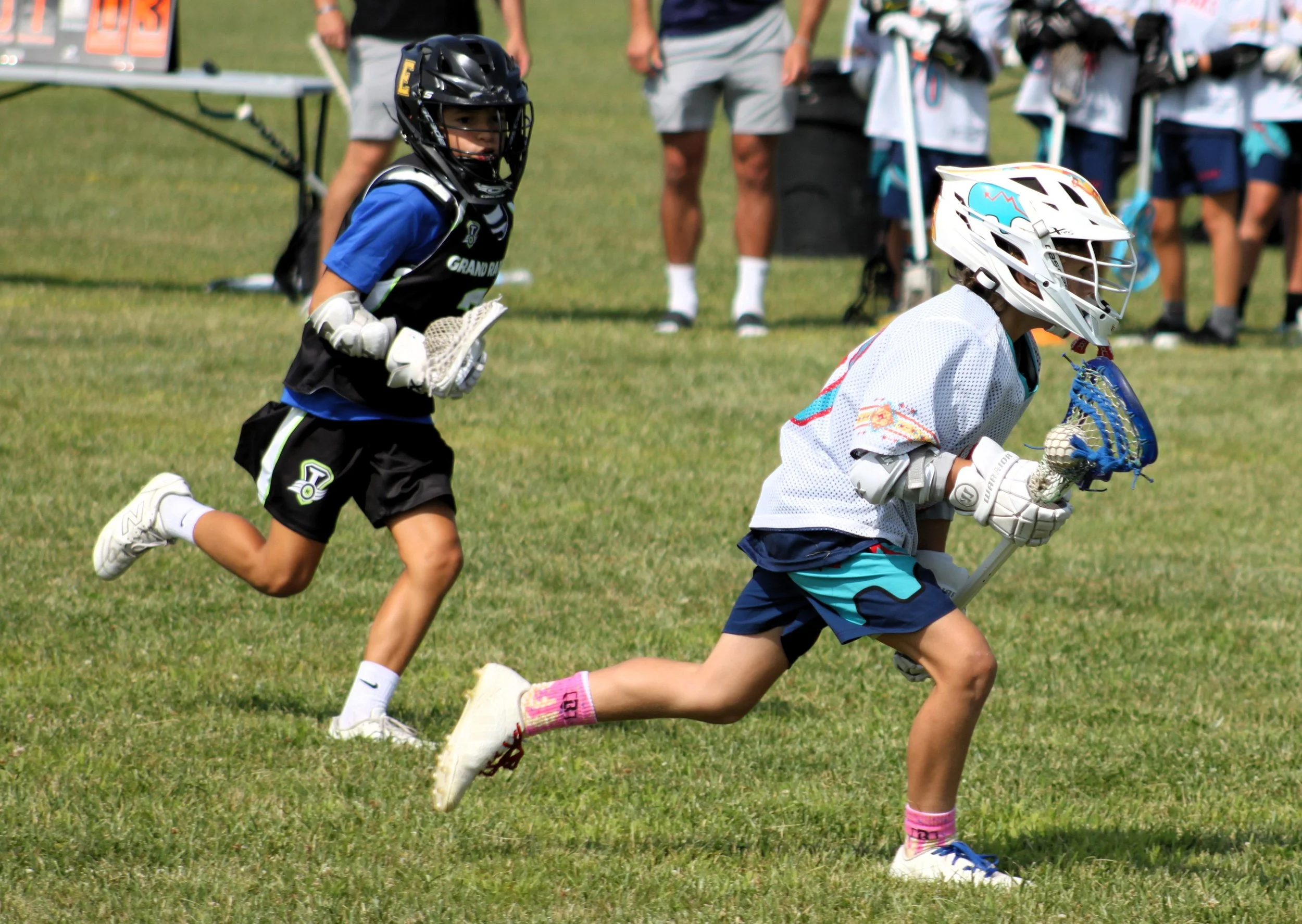 Children playing lacrosse on a grassy field, wearing helmets and sports gear, with other kids and spectators in the background.