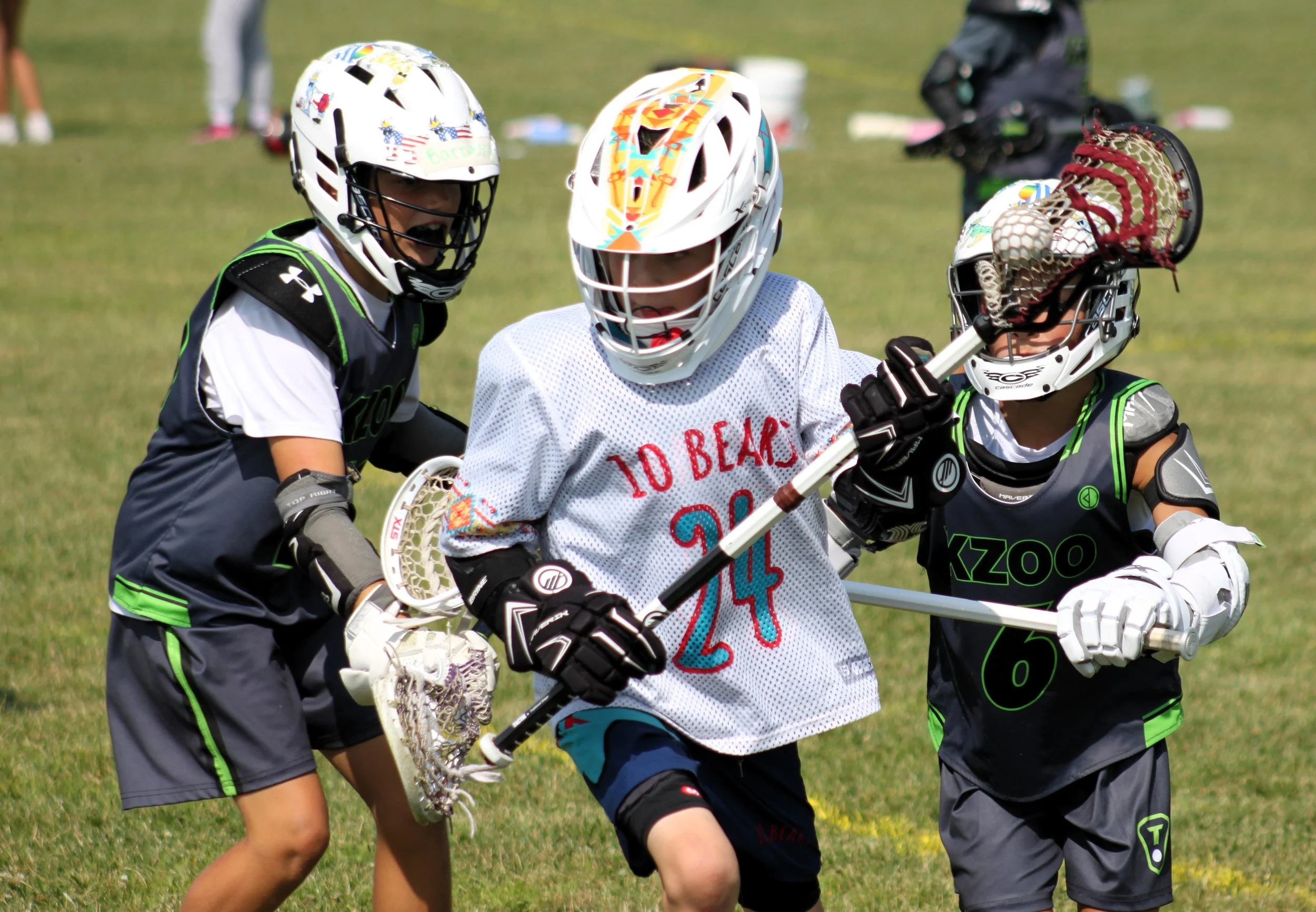 Youth playing lacrosse outdoors, wearing helmets and gear, on a grassy field.