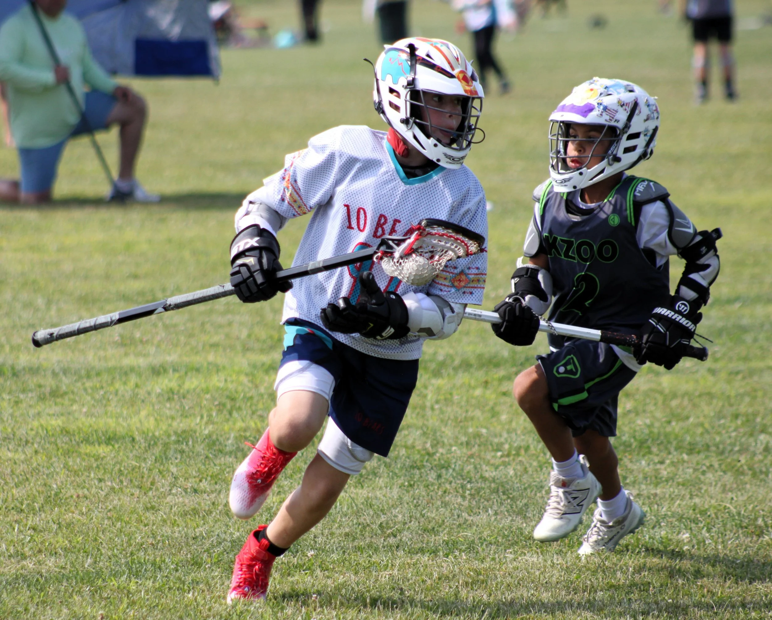 Two children playing lacrosse outdoors on grass, wearing helmets and sports gear, actively engaged in a game.