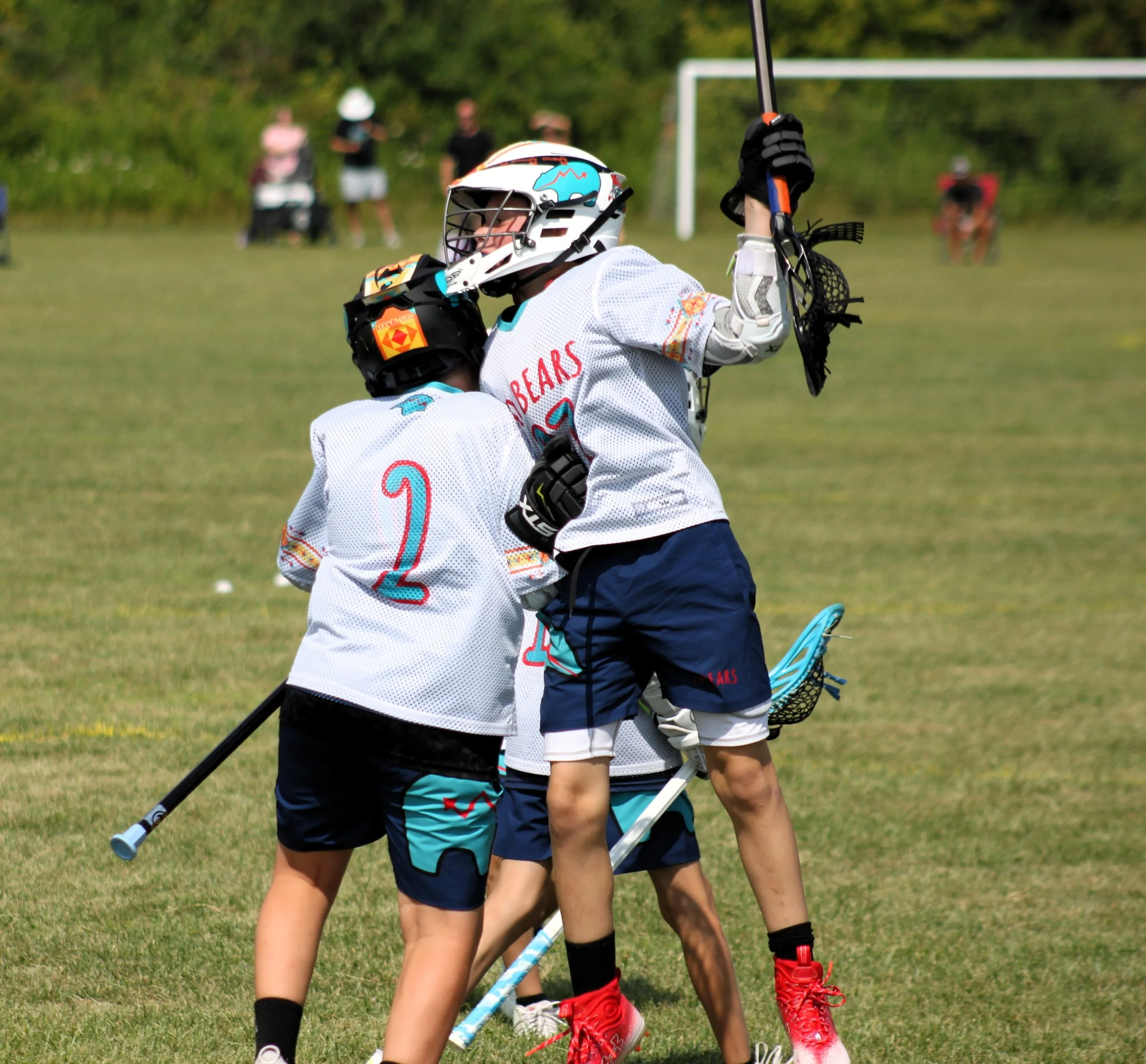 Children playing lacrosse on a grassy field during the daytime, with some spectators in the background.