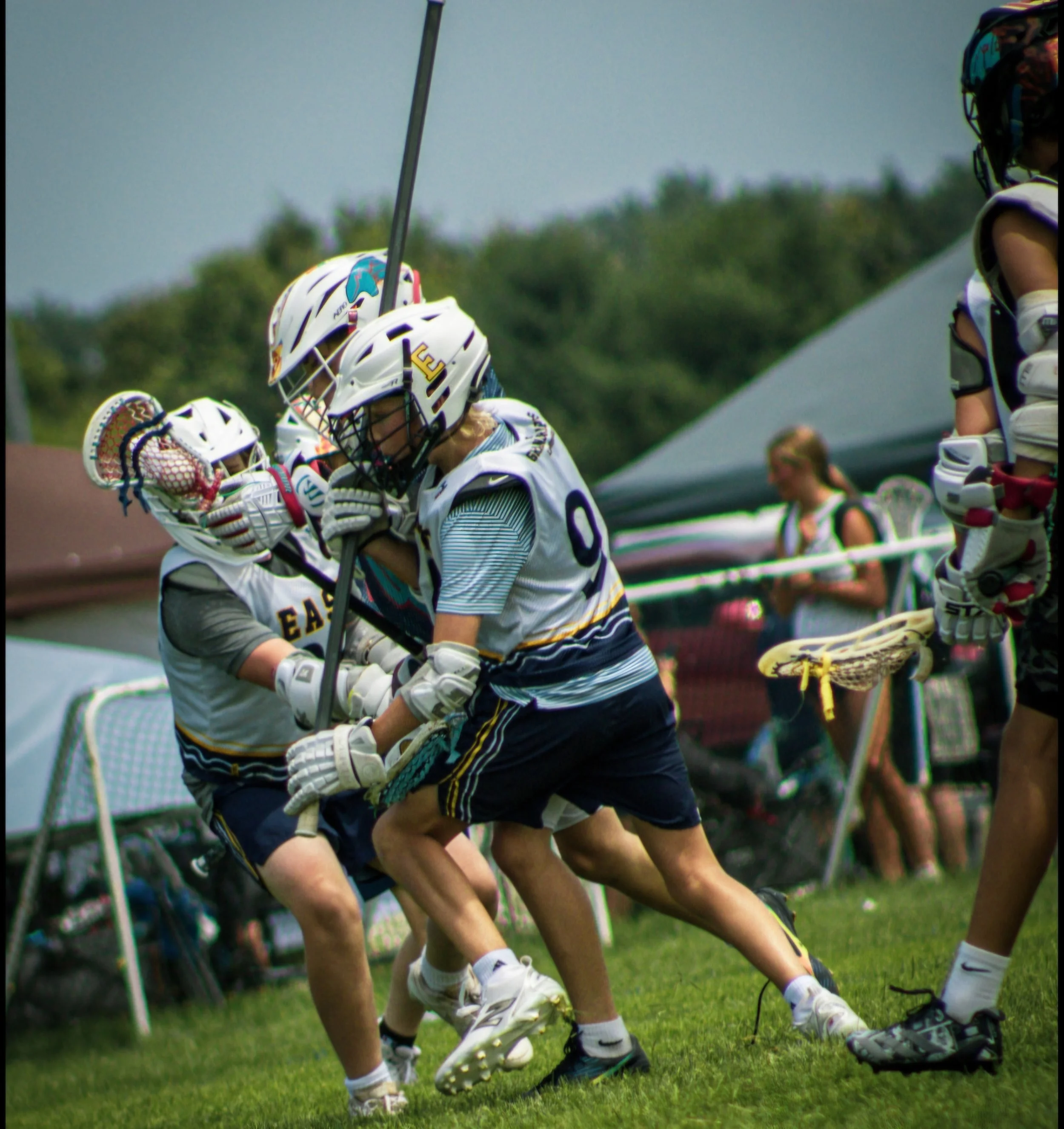 Lacrosse players in white jerseys and helmets compete for the ball near the sidelines on a grassy field, with a line of players and coaches in the background.