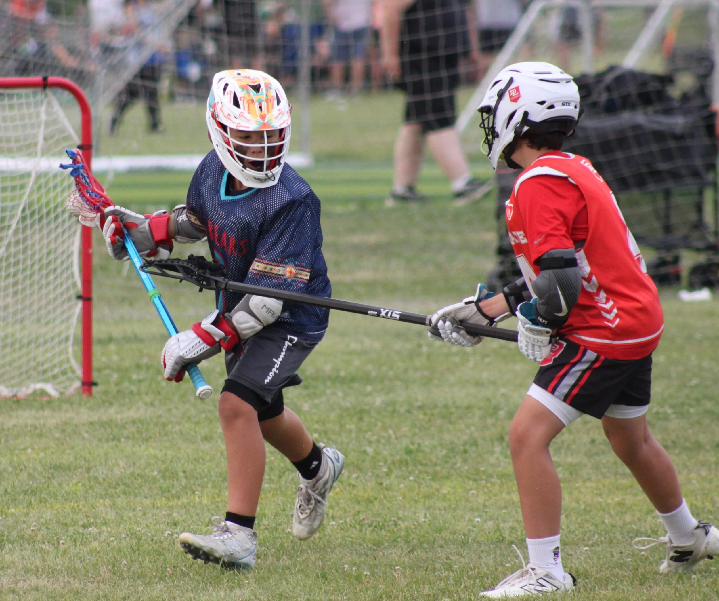 Two young boys playing lacrosse outdoors near the goalpost, both wearing helmets and sports gear, with a netted goal in the background.