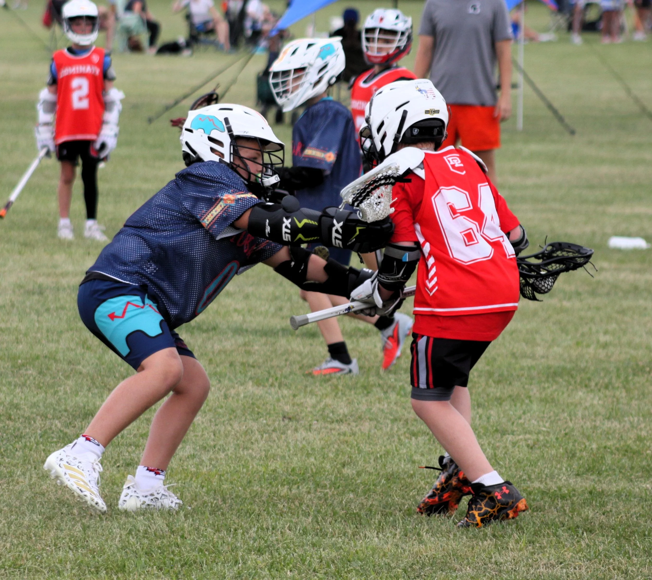 Two young children playing lacrosse on a grassy field, wearing helmets, protective gear, and jerseys, with adults and other children in the background.