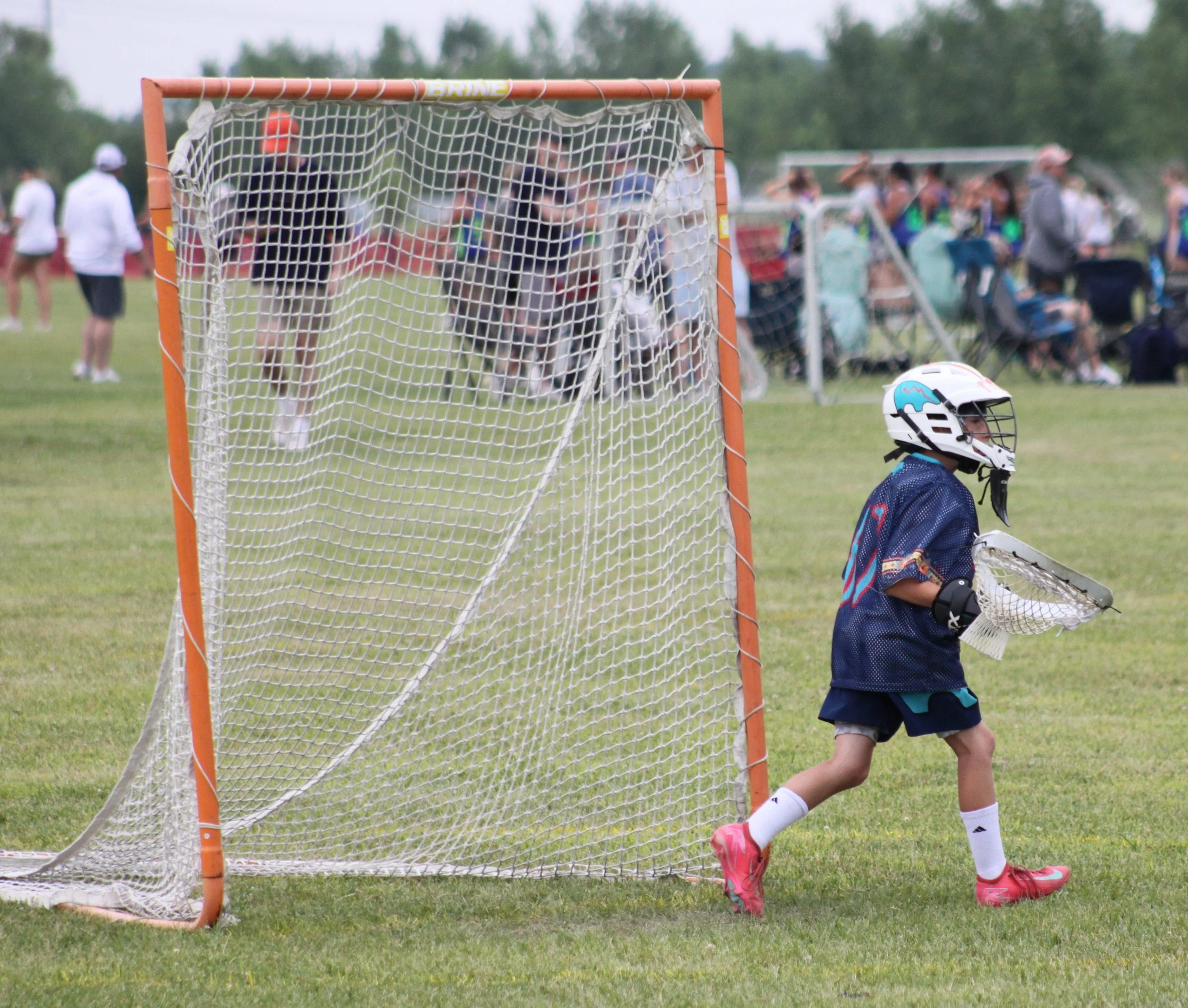 Youth playing lacrosse on a field with spectators in the background, focused on a lacrosse goal with a butterfly-shaped helmet nearby.
