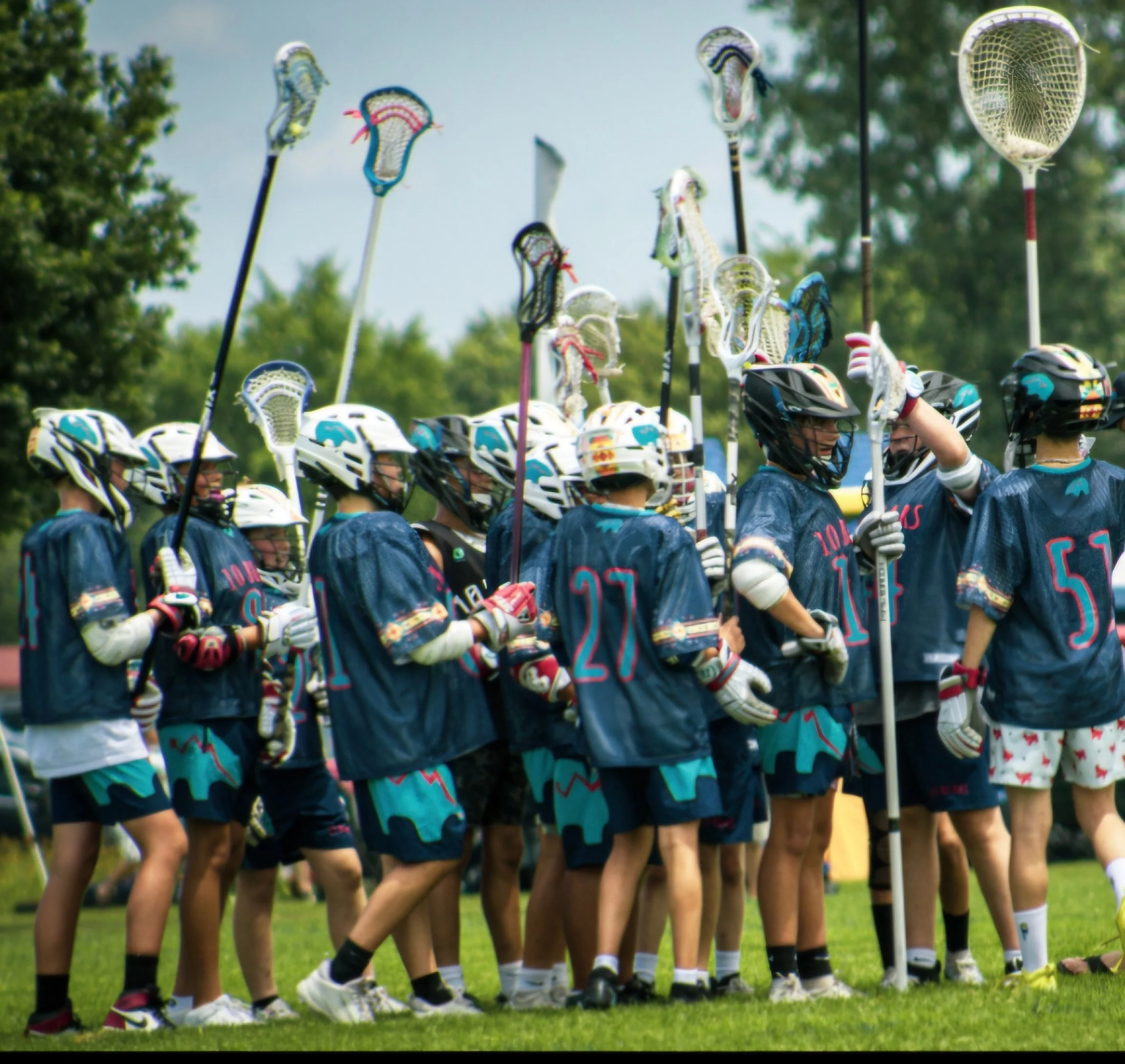 A group of young athletes, wearing matching blue sports uniforms and helmets, standing in a line on a grassy field during daytime, with lacrosse sticks raised vertically in front of them.
