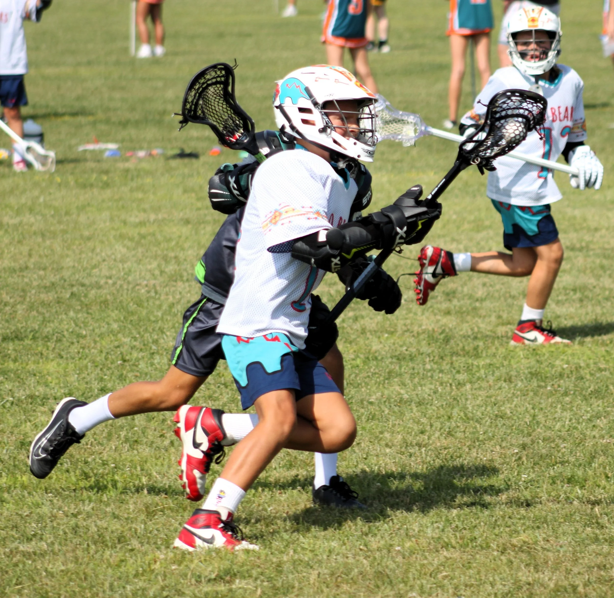 Children playing lacrosse on a grassy field, wearing helmets and sports gear.