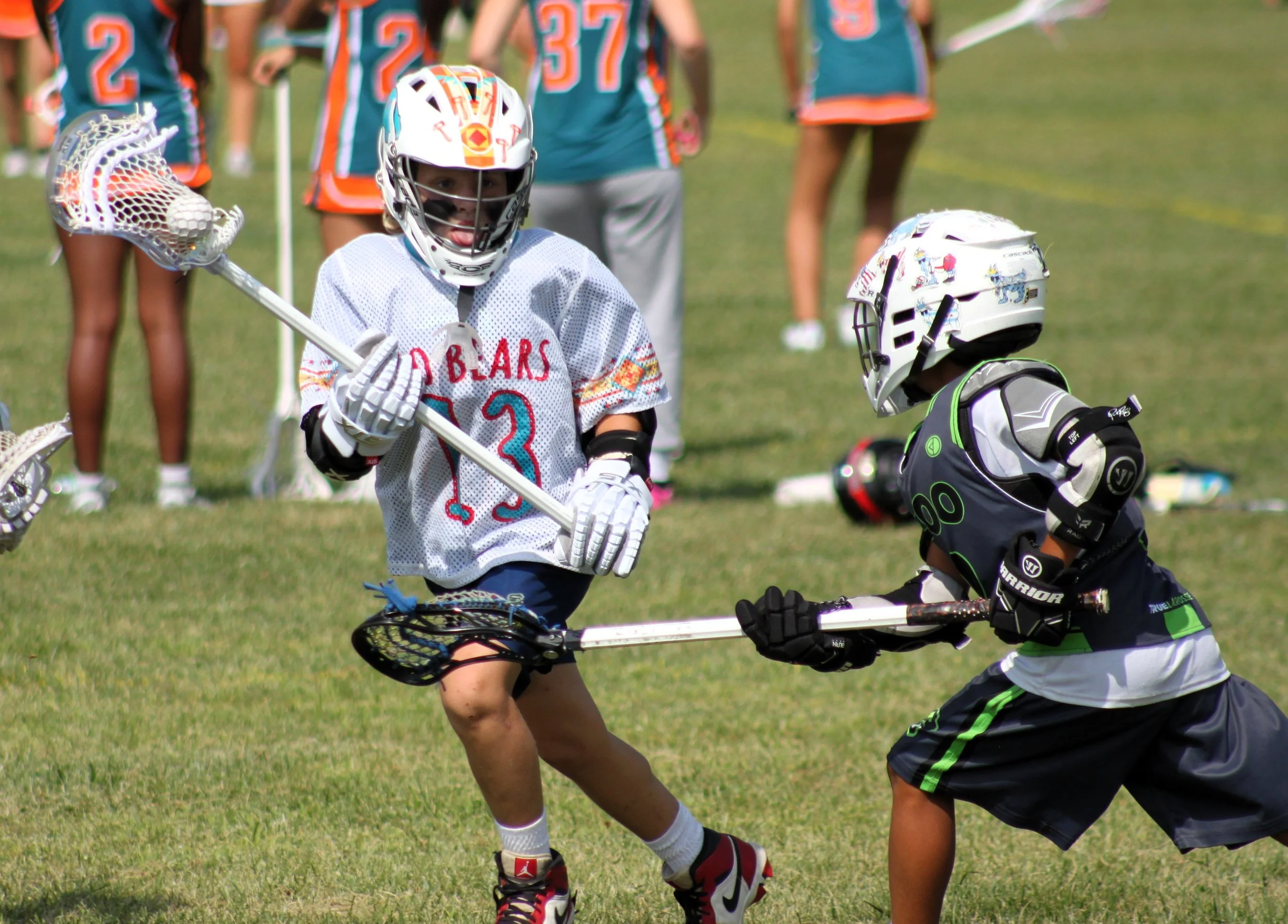 A youth girl playing lacrosse on a grass field, wearing a white jersey with the number 3, a helmet, gloves, and holding a lacrosse stick. Other children are in the background wearing sports uniforms.
