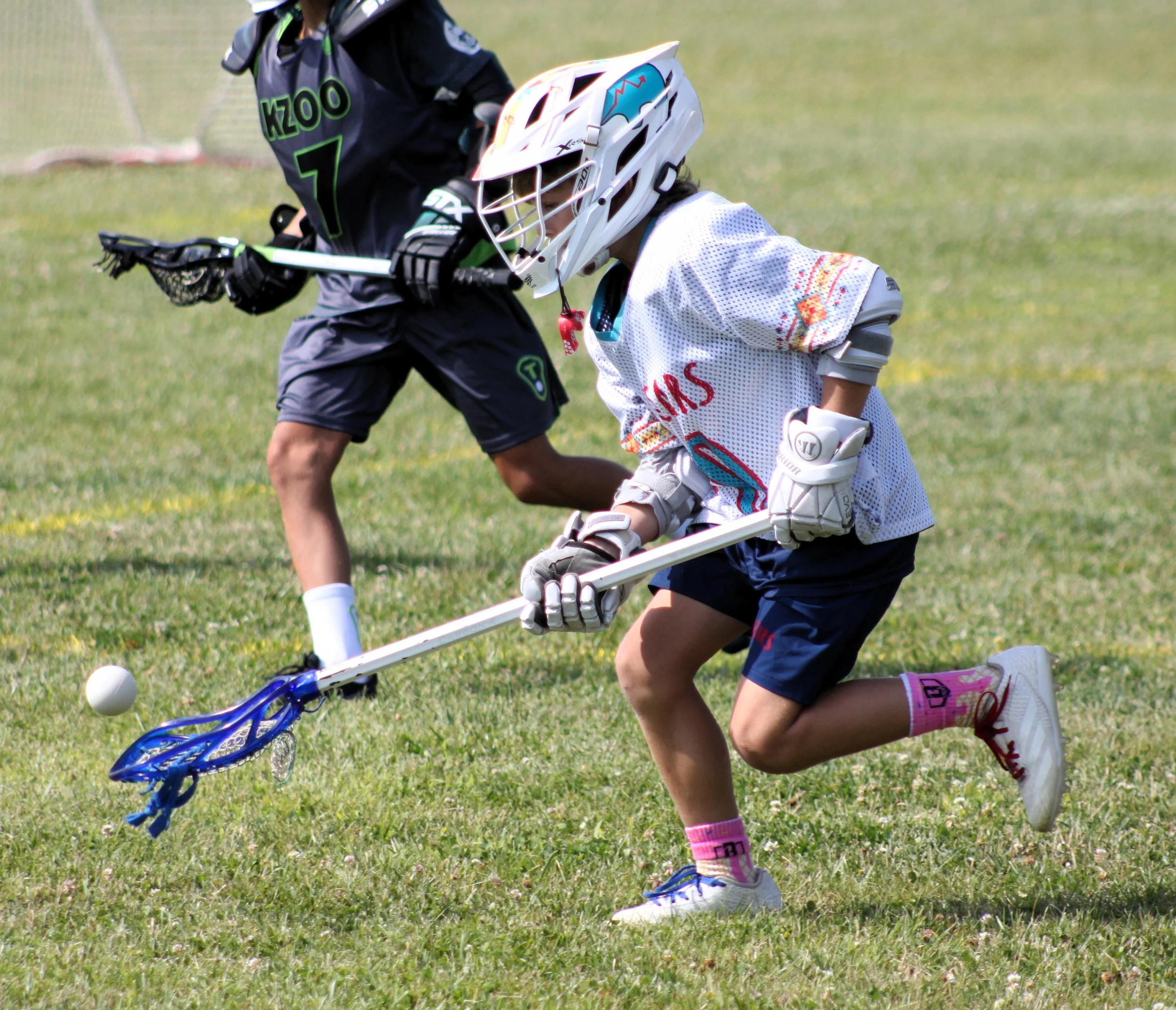 Children playing lacrosse on a grassy field, wearing helmets and sports gear, one child bent down preparing to throw the ball.