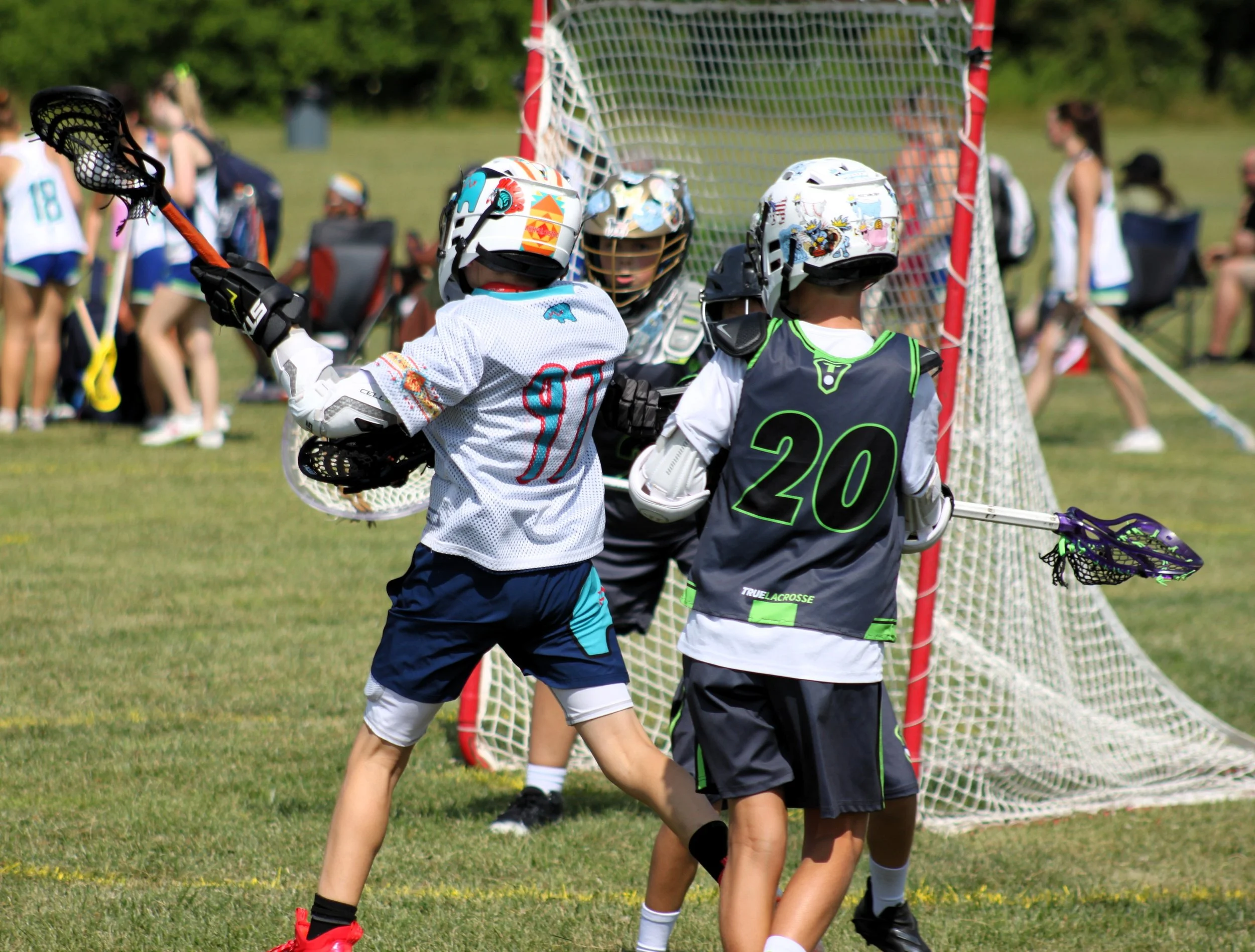 Lacrosse players in protective gear, including helmets and jerseys, gathered near a lacrosse goal during a game on a grassy field with spectators in the background.
