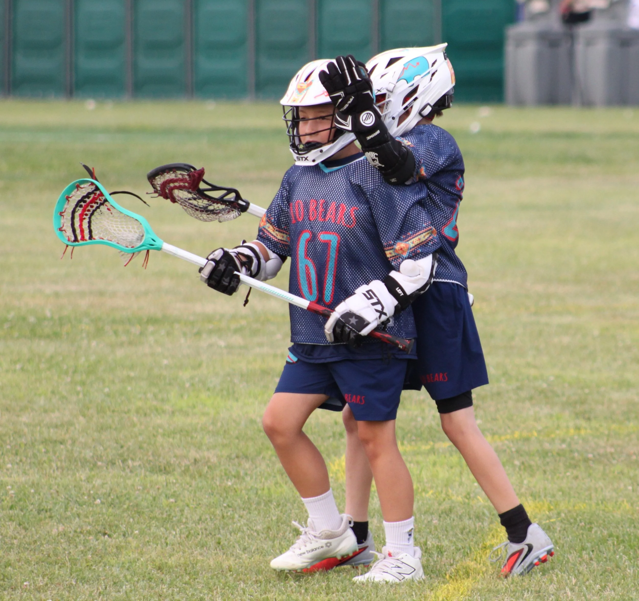 Young children playing lacrosse outdoors on grass field, wearing helmets and sports gear, holding lacrosse sticks.