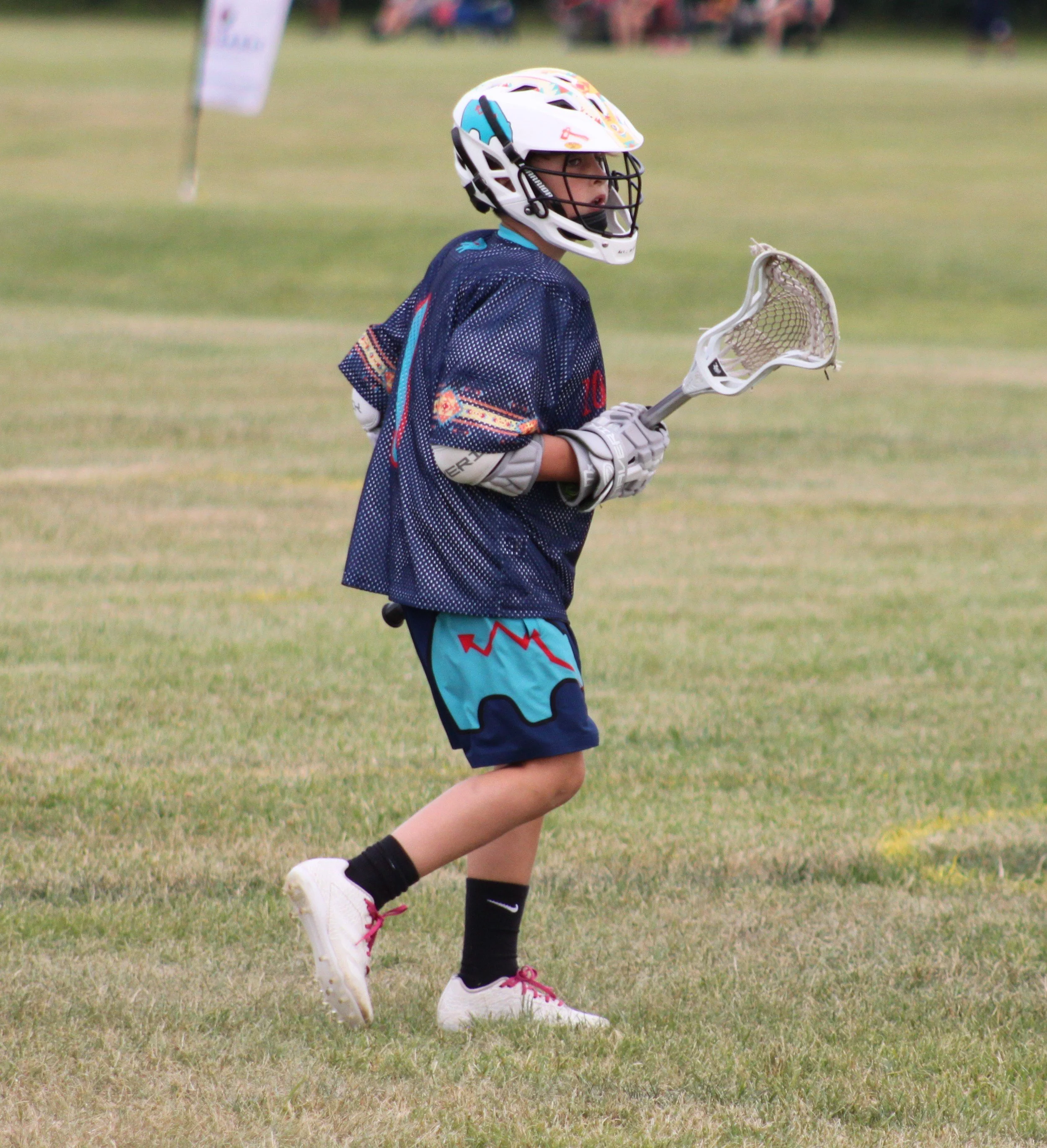 A young boy in lacrosse gear, including a helmet and glove, holding a lacrosse stick on a grassy field.