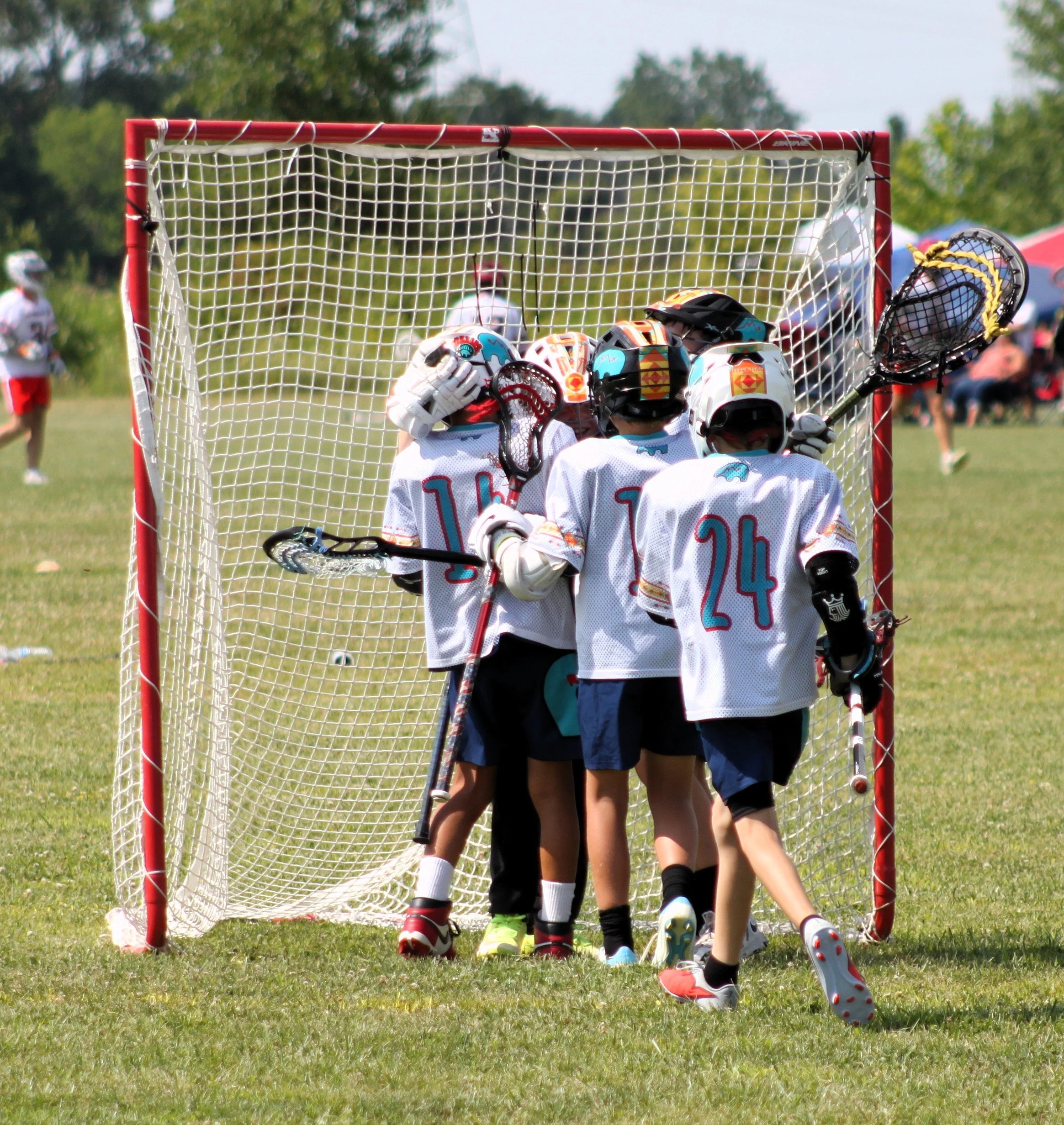 A youth lacrosse team gathers in front of the goal on a grassy field during a game, with players in white jerseys and protective gear.