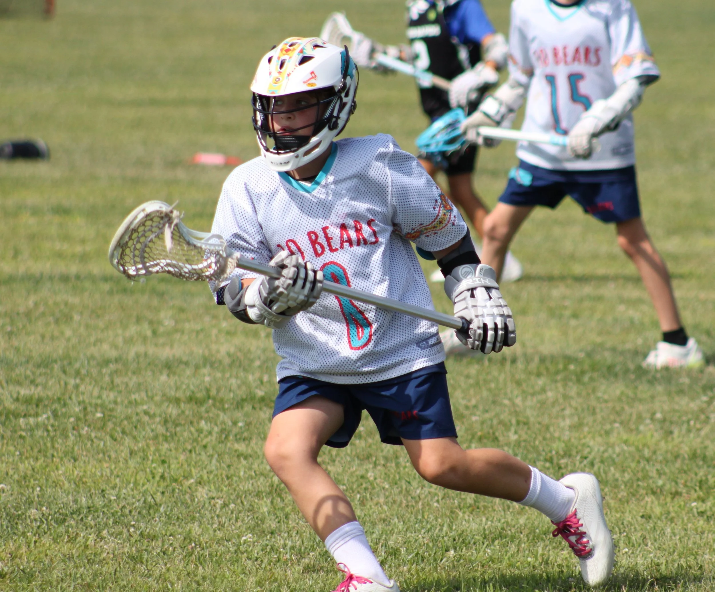 Young boy playing lacrosse on a grassy field, wearing a white jersey with red and blue lettering, a helmet with a colorful design, gloves, and holding a lacrosse stick.