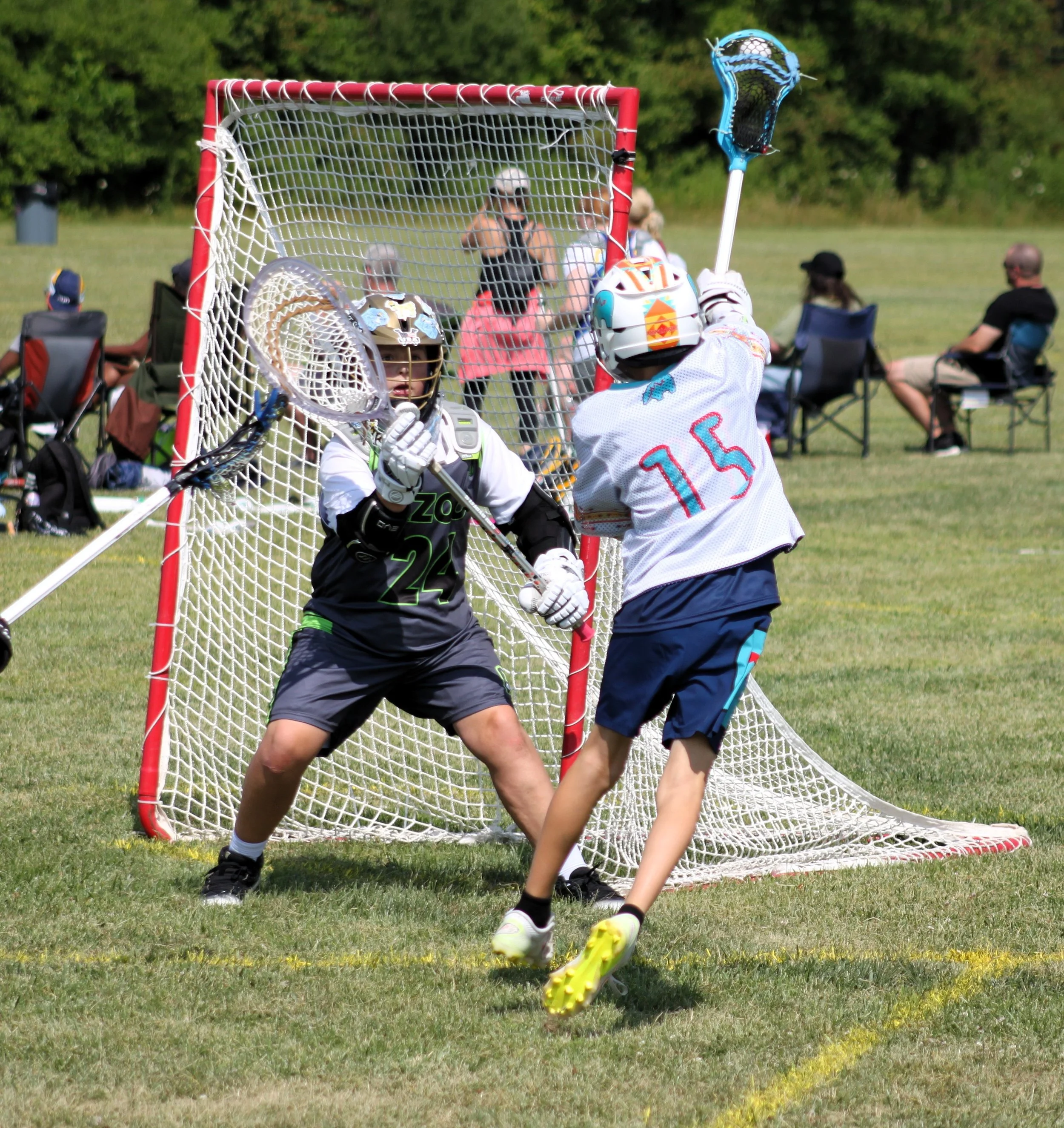 Children playing lacrosse on a grassy field, with spectators sitting in chairs in the background.