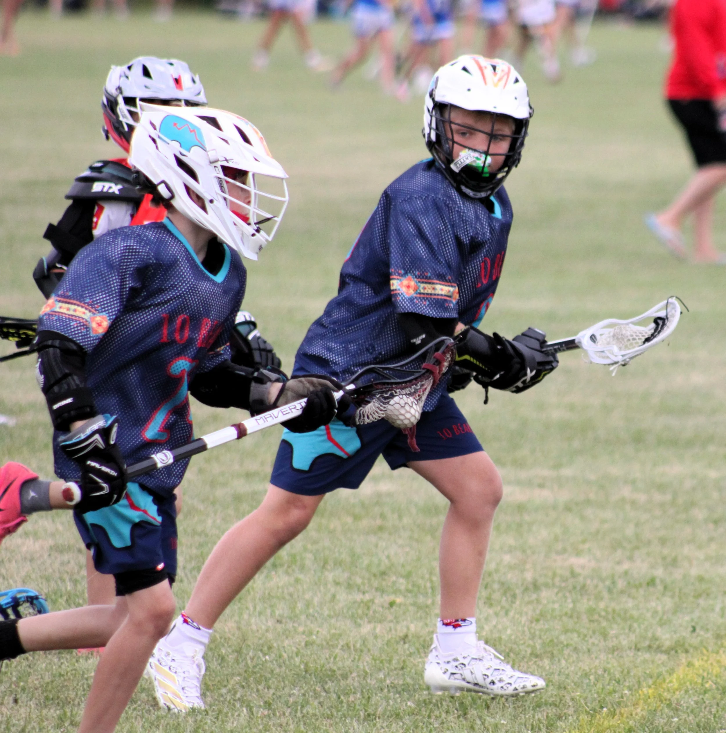 Children playing lacrosse on a grassy field, wearing protective helmets and sports uniforms, with other players in the background