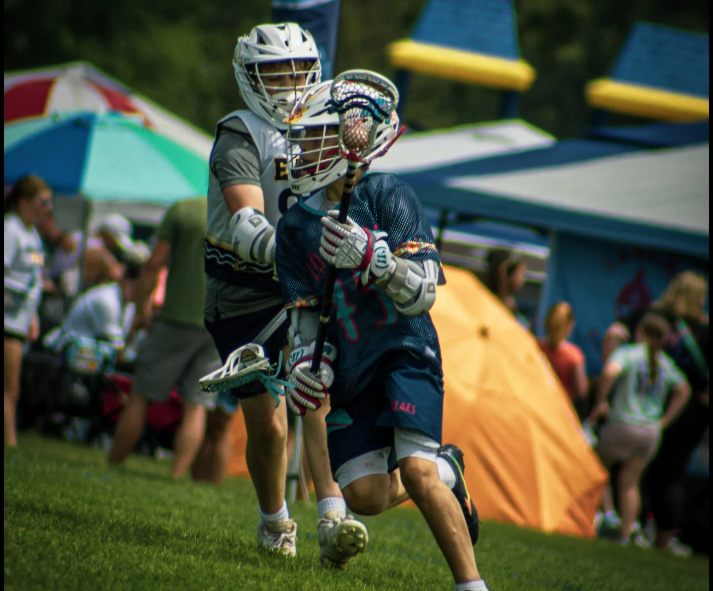 A youth playing lacrosse, wearing protective gear including a helmet, goggles, gloves, and holding a lacrosse stick, during a game or practice session on a grassy field with colorful umbrellas and other players in the background.