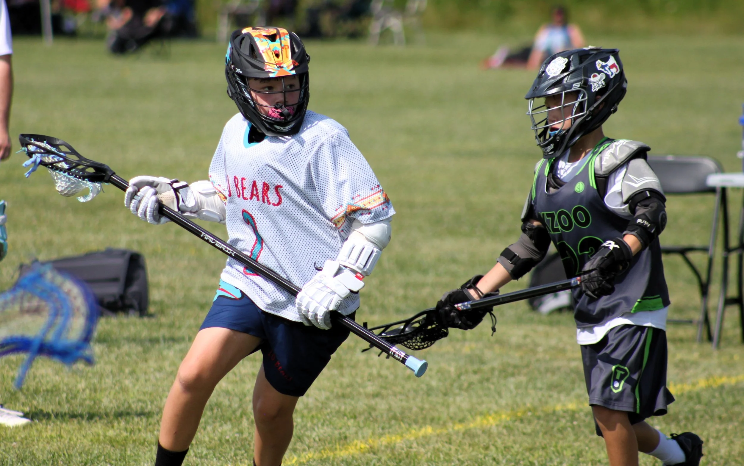 Two children wearing hockey gear playing lacrosse on a grassy field.