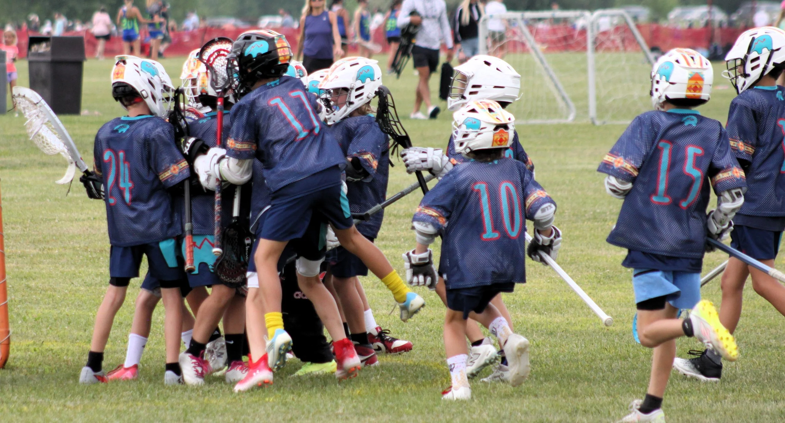 Children in blue sports uniforms with helmets and sticks, playing lacrosse on a grassy field during a game or practice.