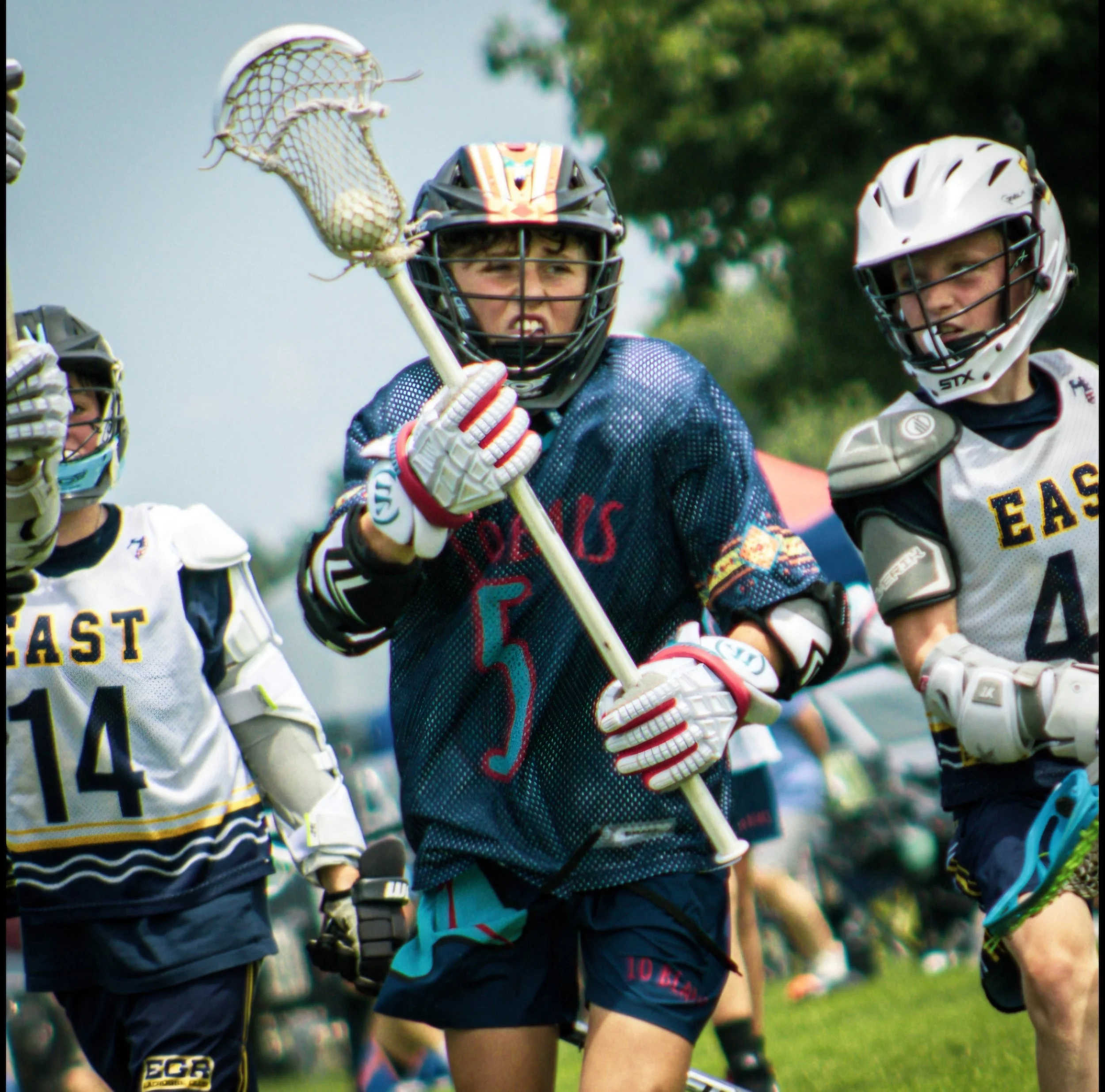 Youth hockey players in uniforms and helmets, one holding a hockey stick and the other with a lacrosse stick, on a grassy field with trees in the background.
