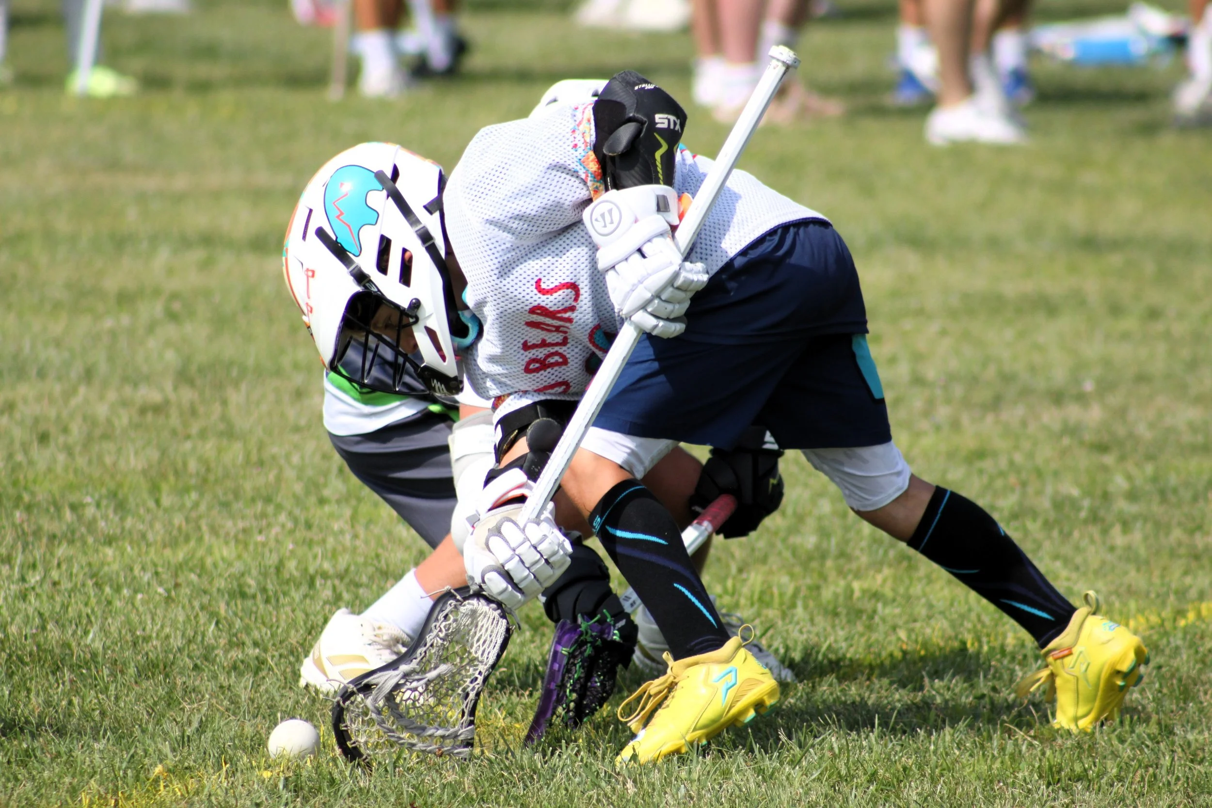 Two young boys playing field hockey on a grassy field, wearing helmets and sports gear, with one boy in white jersey and the other in black shorts, both engaged in a game.