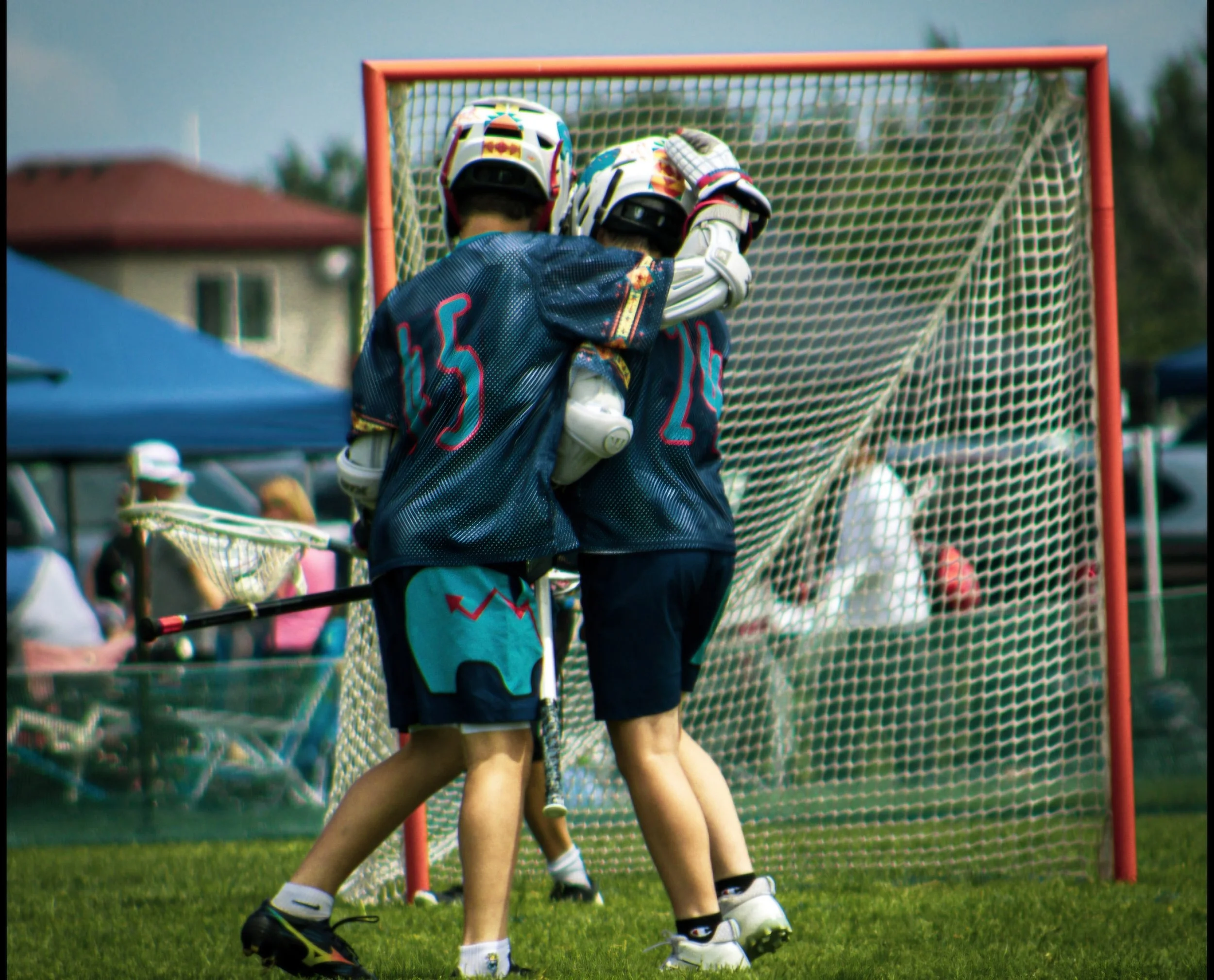 Two young lacrosse players wearing helmets, gloves, and jerseys with the number 55, engaged in a game near the goal net.