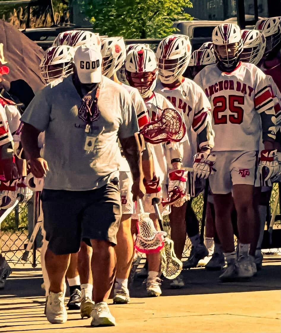 A lacrosse team walking with their coach on a sideline, wearing white uniforms with red accents, and helmets, some with face shields, during daytime.
