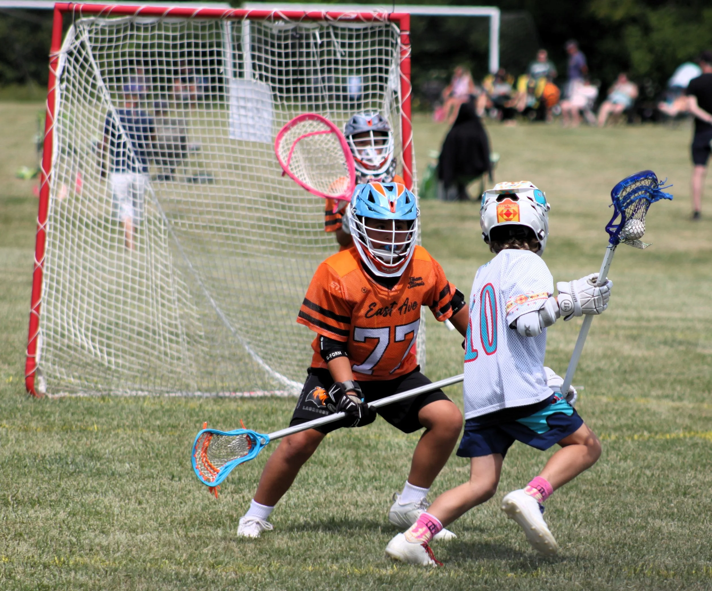 Youth lacrosse game with children playing, one colorful goalie in orange and blue gear in front of the goal, others competing for the ball, spectators in the background.
