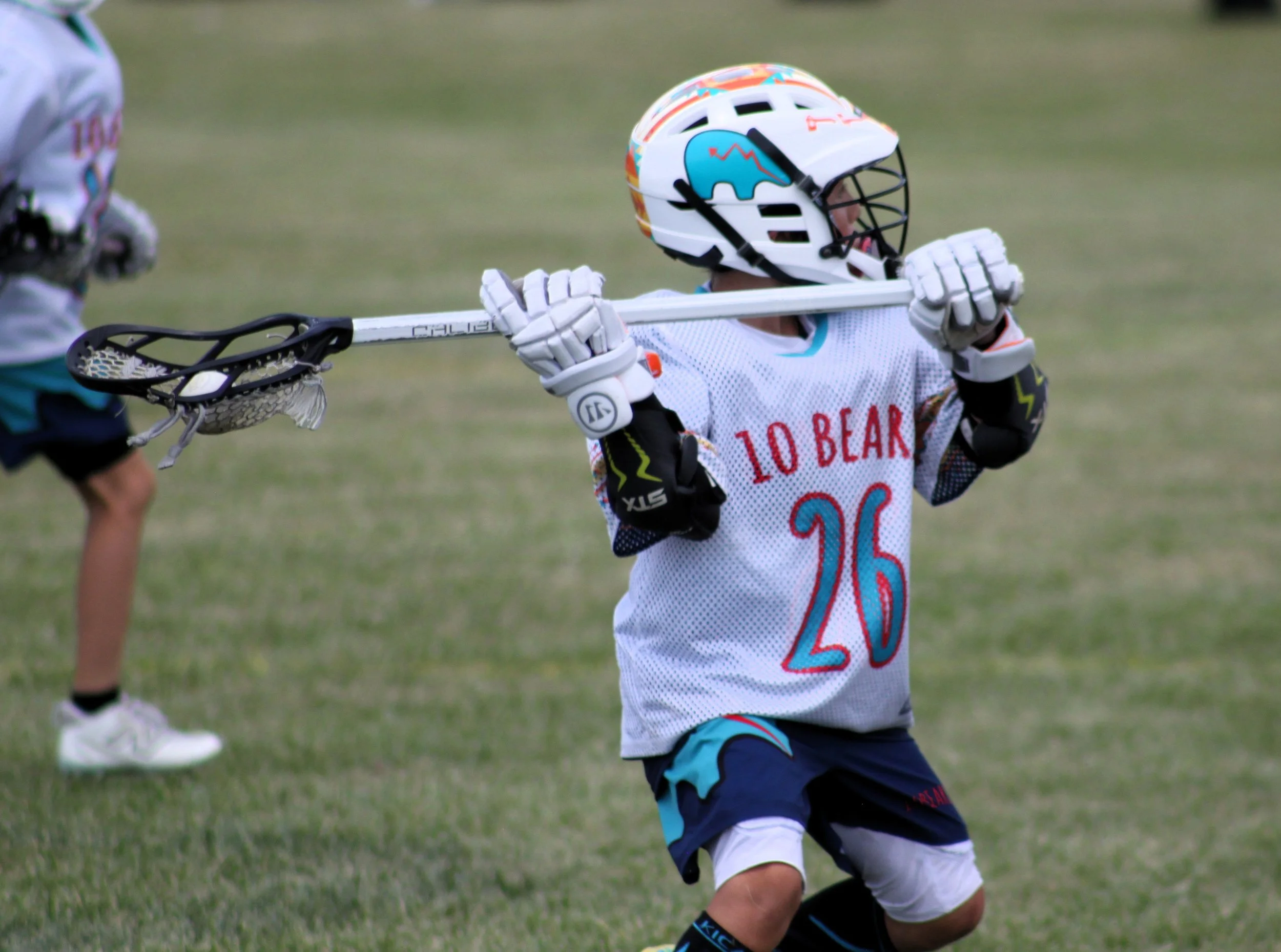 A young lacrosse player wearing a white jersey with red and blue details, black and blue shorts, a white helmet with a colorful design, and wearing gloves, is running on a grassy field while holding a lacrosse stick.