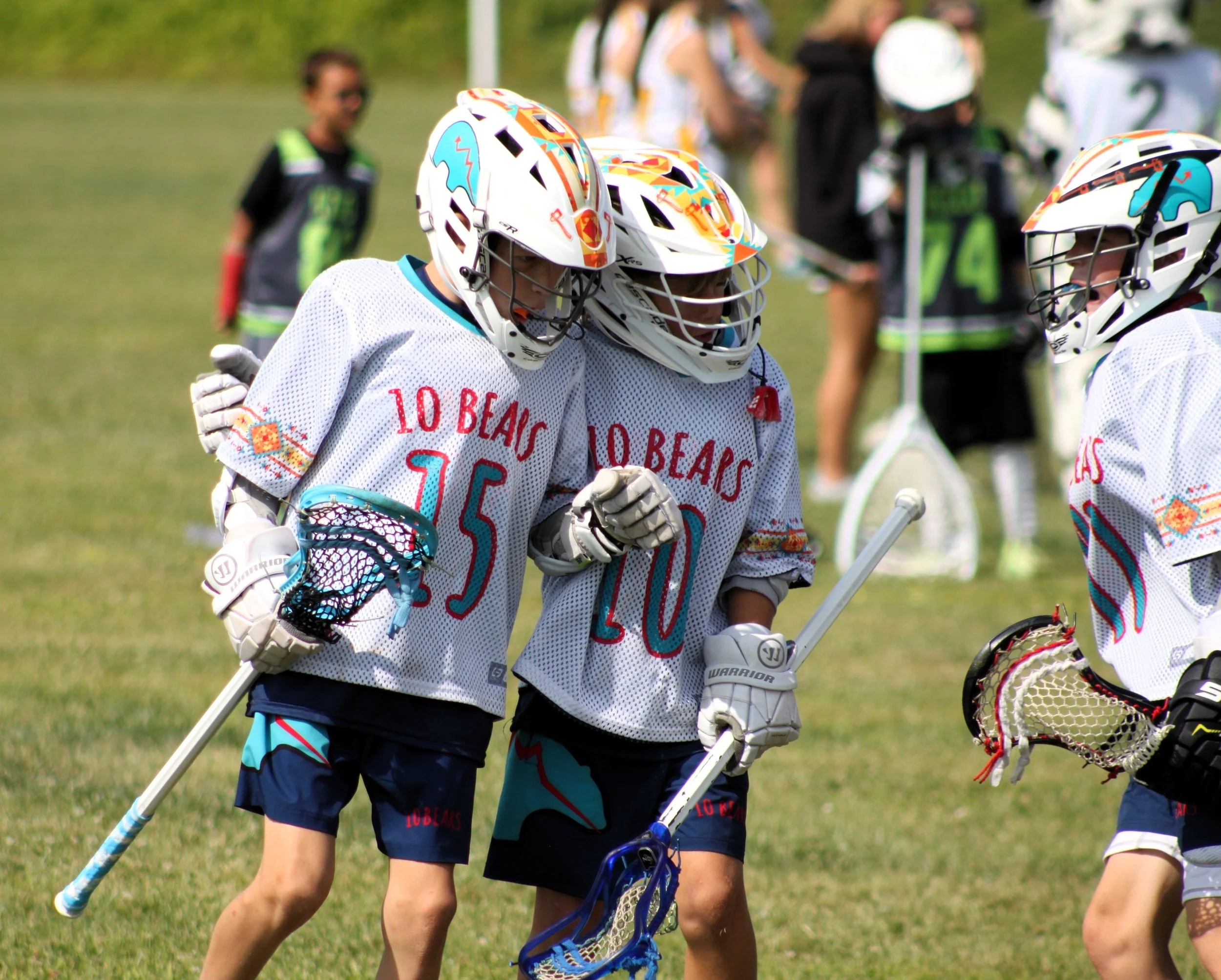 Lacrosse players in white jerseys with colorful designs huddle together on a grassy field during a game, with other players and spectators in the background.