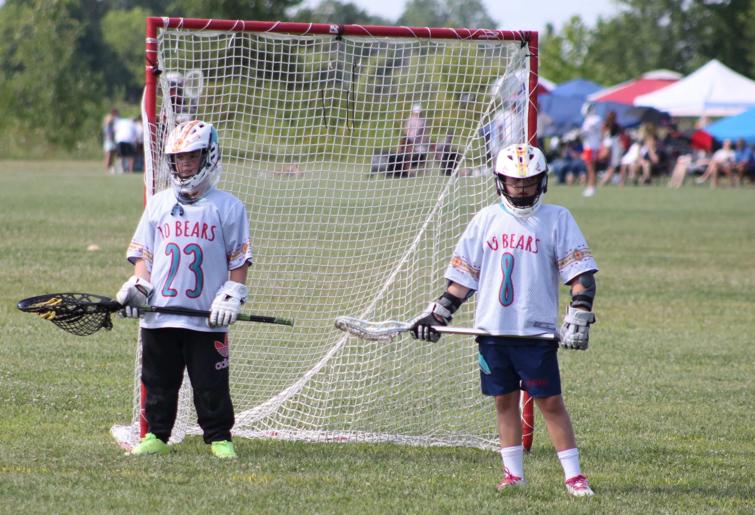 Two young boys dressed in hockey gear standing next to a hockey goal on a grassy field during a sporting event. The boys are wearing helmets and jerseys, with one boy holding a hockey stick. The background shows people sitting under tents and umbrell