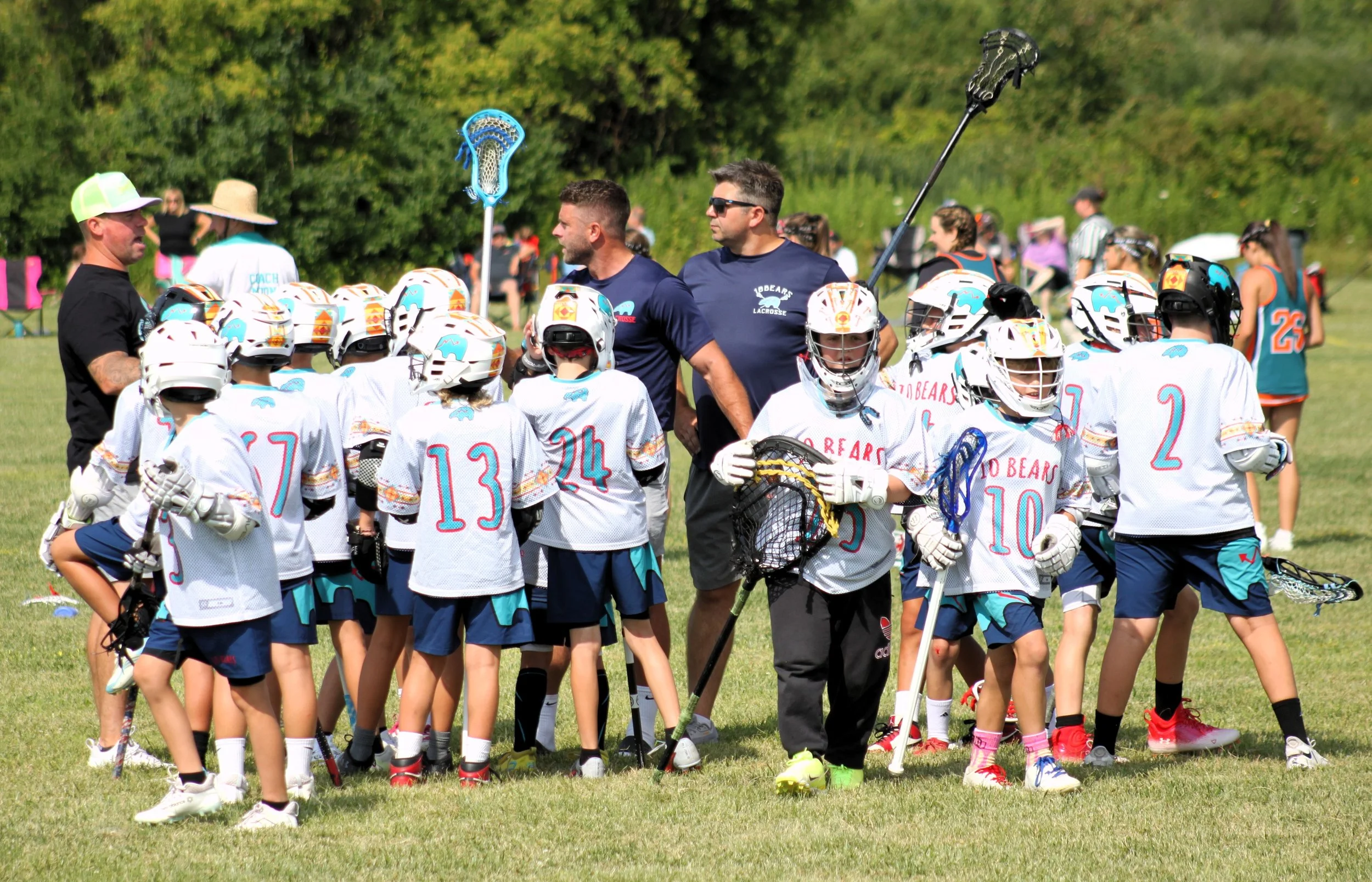 A group of young lacrosse players in white jerseys and helmets gather around their coaches on a grassy field during a daytime practice or game. The coaches are talking to the team, with one holding a lacrosse stick and another with sunglasses. Other 