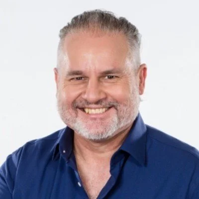 Headshot of a middle-aged man with gray hair and beard, wearing a blue collared shirt, smiling against a plain light background.