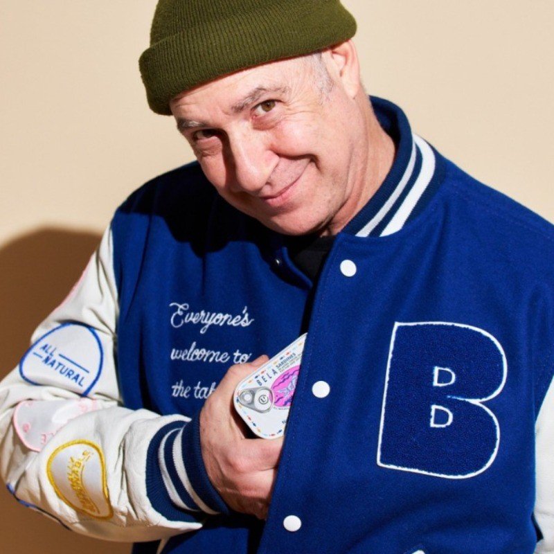 A smiling man wearing a green beanie and a blue varsity jacket with patches, holding a deck of playing cards.