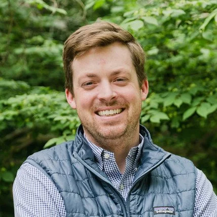 A man smiling outdoors, wearing a blue vest and a checkered shirt, with greenery in the background.