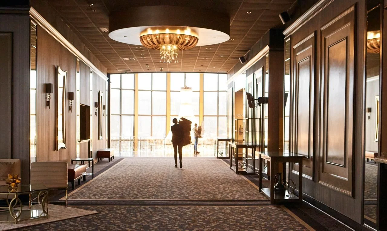 Interior of a modern hotel lobby with wooden paneling, large window, chandelier, and a person walking toward the exit.