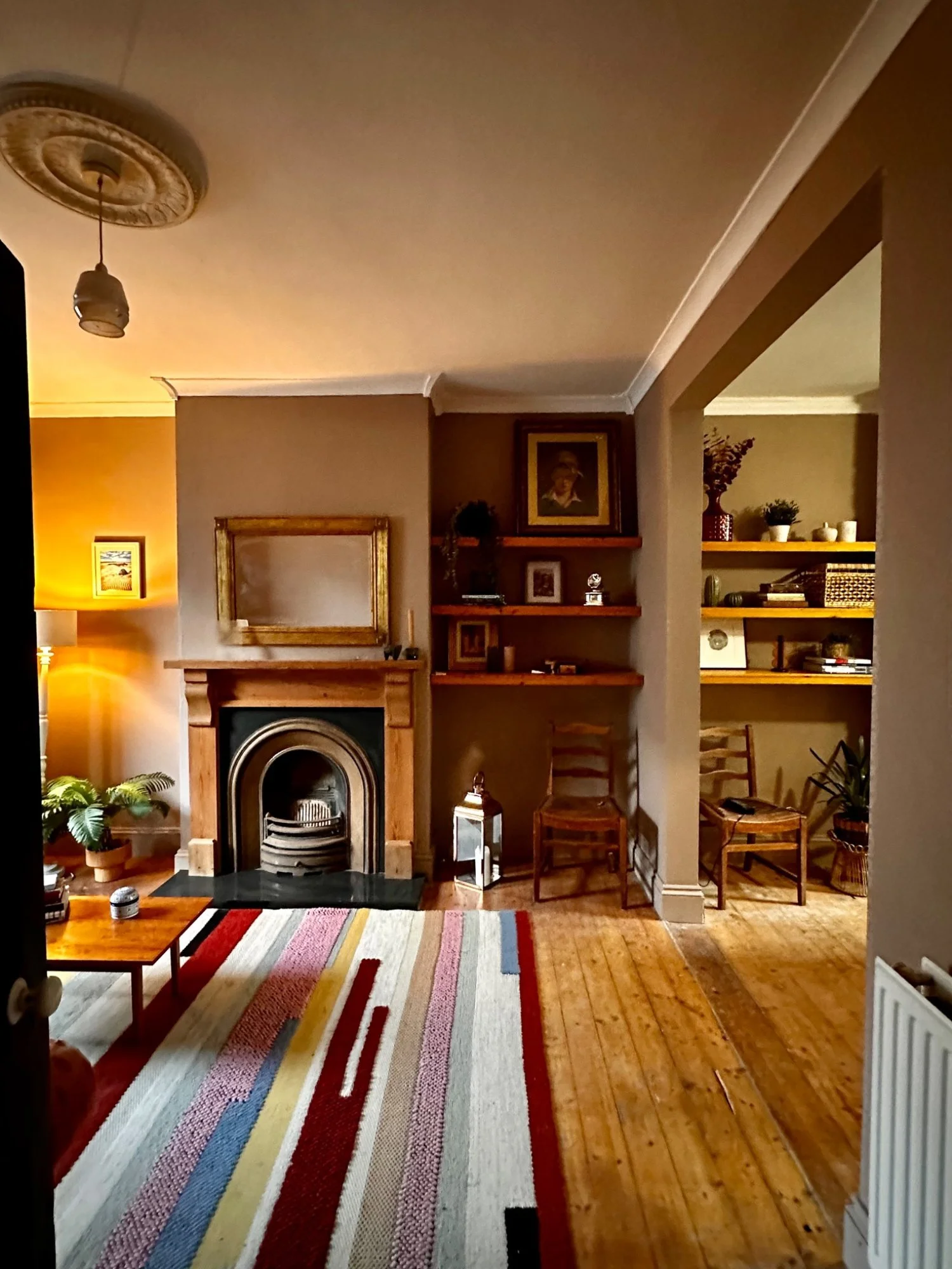 A cozy living room with a wooden fireplace, a colorful striped rug, and built-in wooden shelves with decorations and books, along with a plant and chairs.