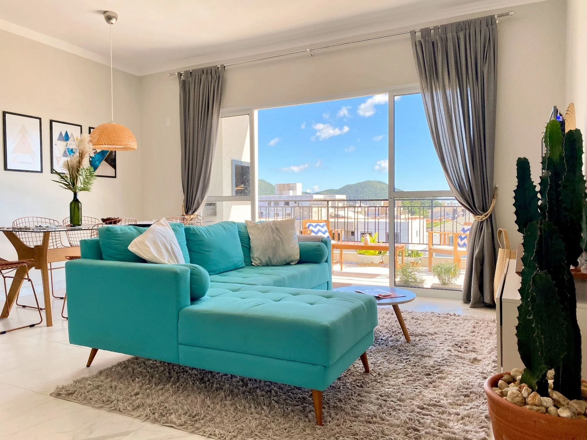 Bright living room with teal sectional sofa, white pillows, beige rug, wooden coffee table, large window with gray curtains overlooking a balcony with outdoor furniture and a mountain view, decorative artwork on the wall, large cactus plant in a terracotta pot.
