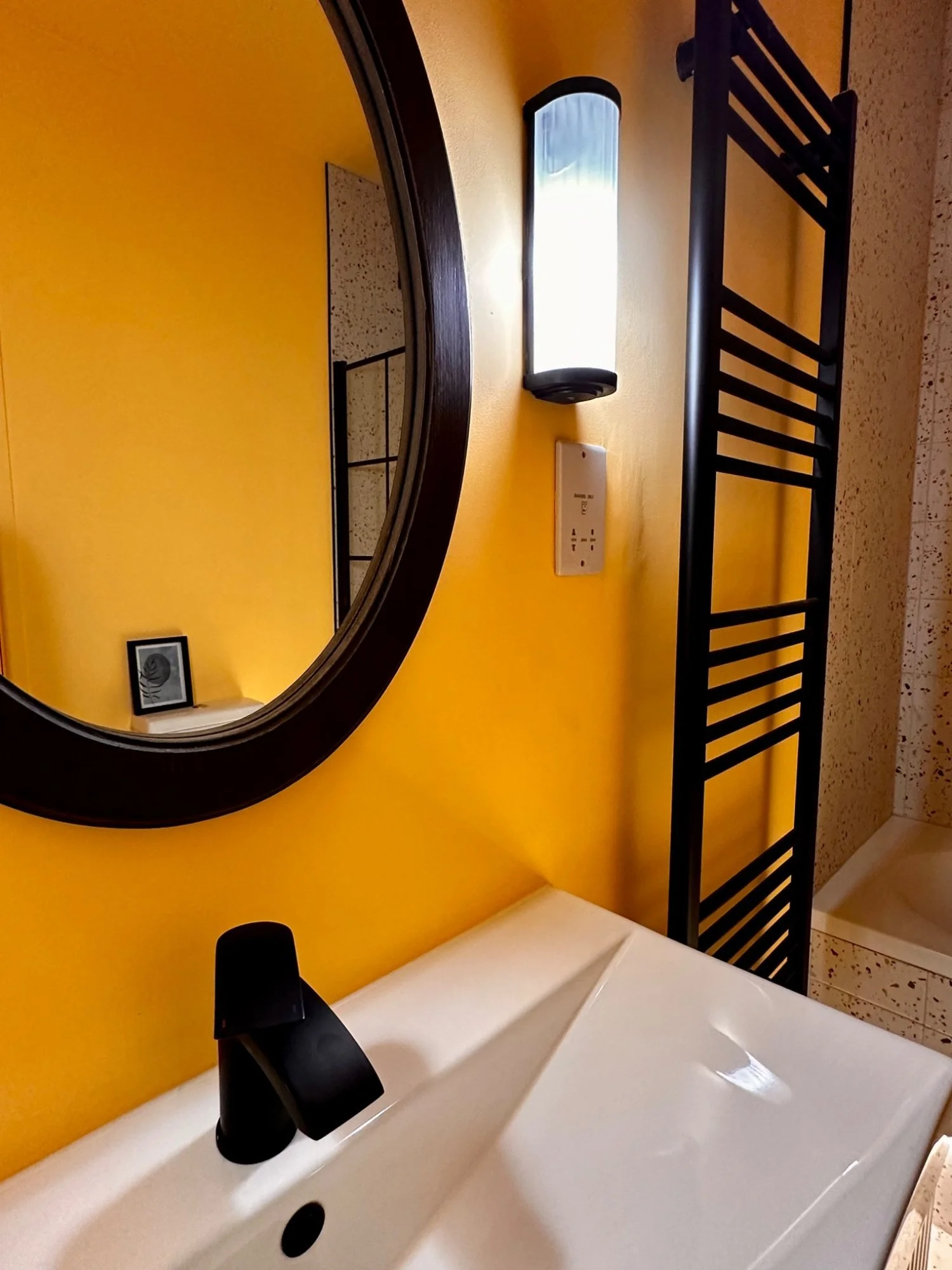Bathroom with yellow wall, oval mirror, black sink faucet, wall light, black ladder towel rack, and shower area in the corner.