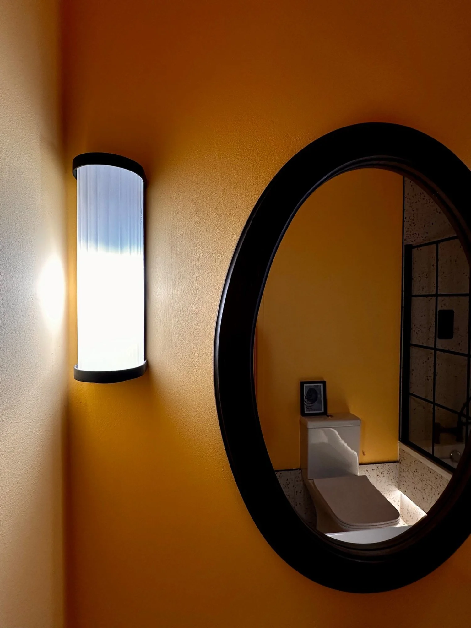 A yellow bathroom wall with a black-framed oval mirror reflecting a white toilet with a faceplate and a beige seat. A tall wall-mounted light fixture illuminates the scene from the left.
